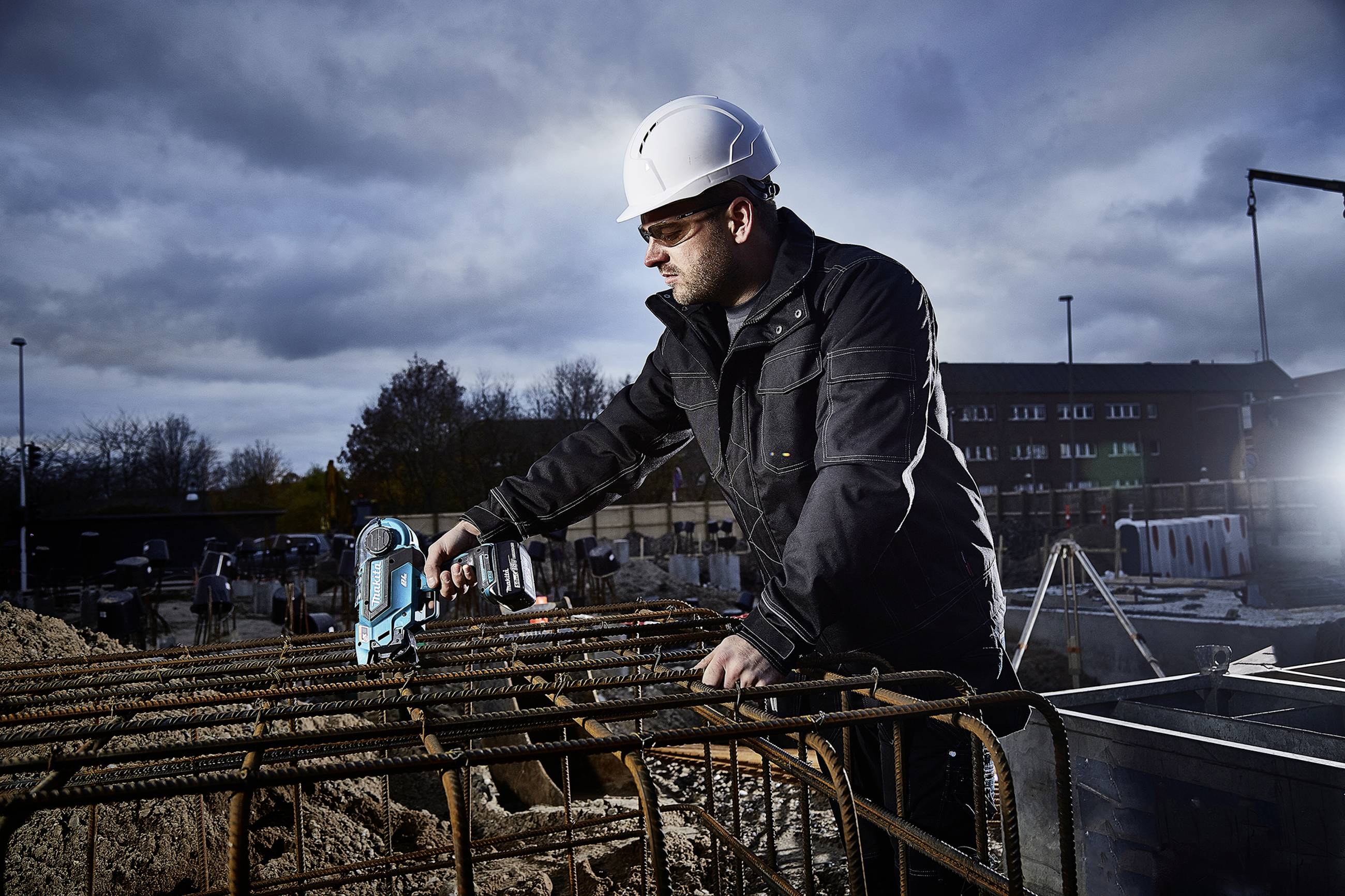 Arbeiter mit Schutzhelm befestigt Stahlstangen auf Baustelle unter bewölktem Himmel. Wichtiger Sicherheitsaspekt im Bauwesen.