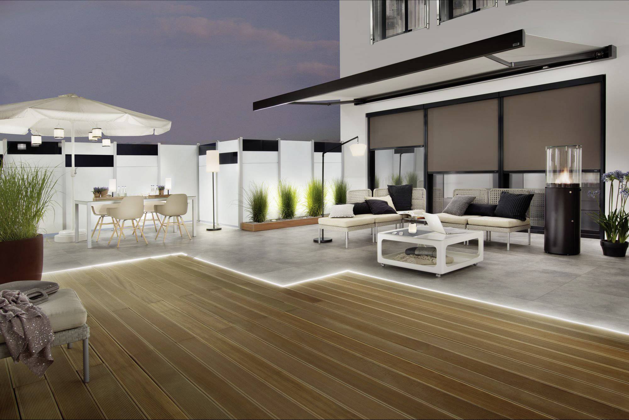 Terrasse bei Nacht mit modernen Möbeln: Sofas, Tisch, Stühle, Pflanzen in Töpfen, Sonnenschirm, stimmungsvolle Beleuchtung.