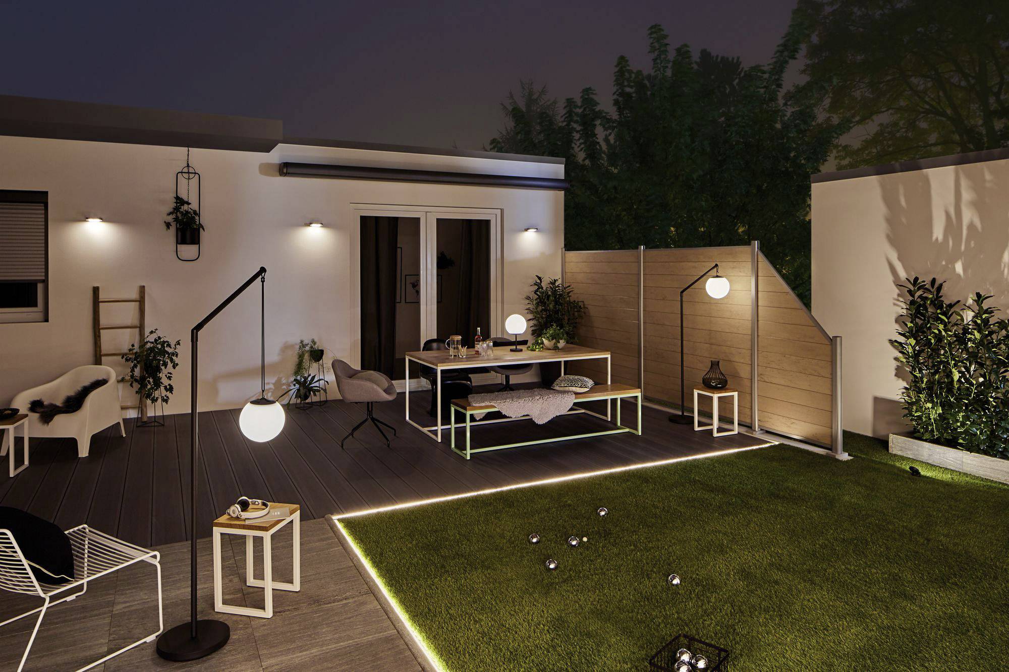 Ein moderner Garten im Abendlicht mit Sitzbereich, Tisch, Pflanzen und Beleuchtung. Im Hintergrund ist ein beleuchtetes Haus zu sehen.