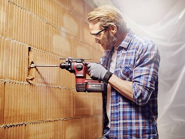 Ein Mann mit Schutzbrille und Handschuhen bohrt mit einer roten Bohrmaschine ein Loch in eine Ziegelwand.