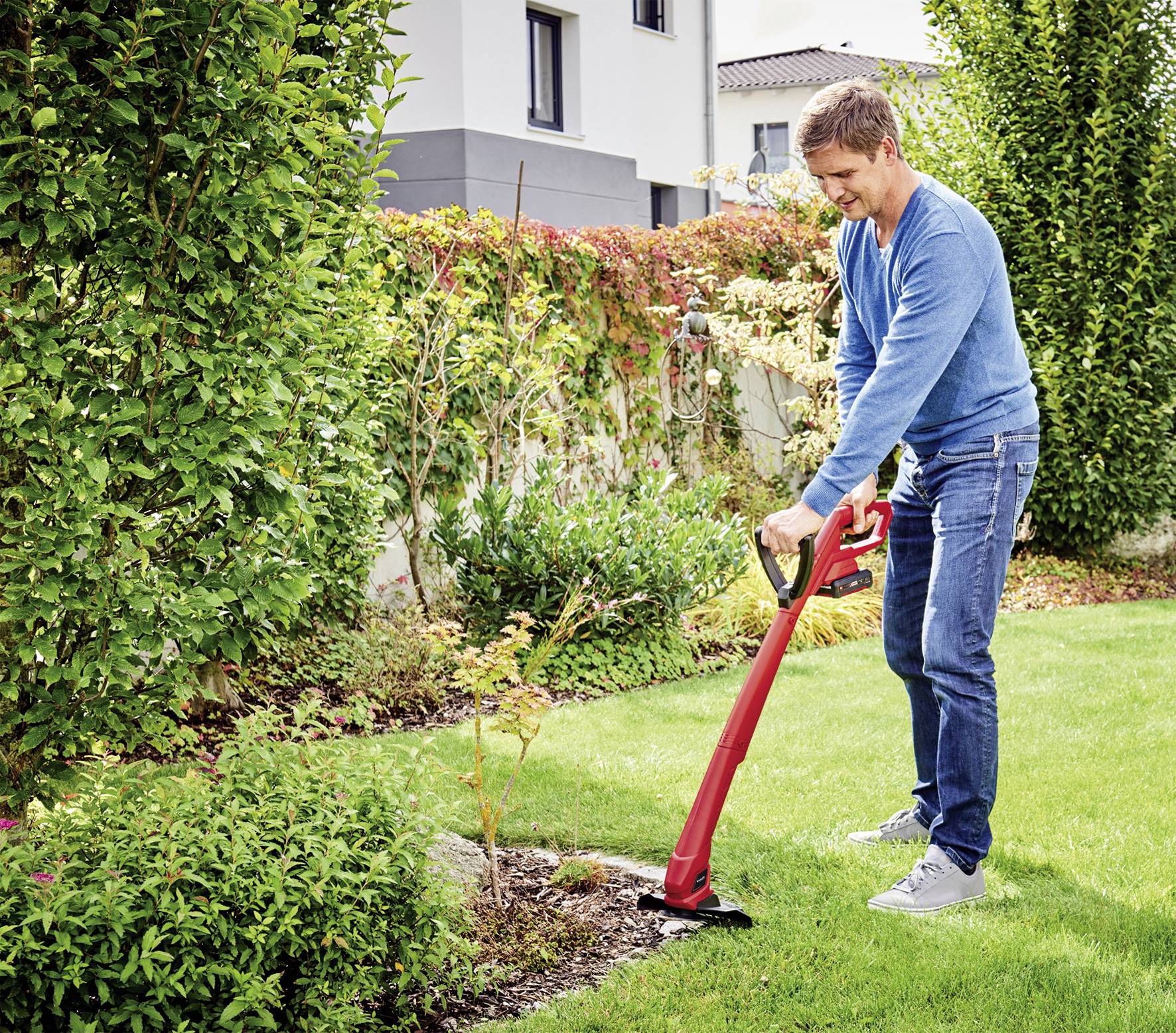 Ein Mann mäht den Rasen mit einem roten Rasentrimmer in einem gepflegten Garten neben einem Haus.