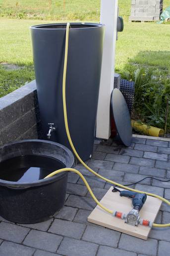 Wasserfass auf einer Terrasse mit gelbem Schlauch, der in einen Eimer führt. Eine Pumpe liegt daneben, bereit zur Bewässerung.