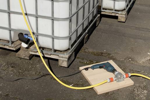 Eine Pumpe pumpt Flüssigkeit über einen gelben Schlauch aus einem großen Wassertank. Die Pumpe liegt auf einem Holzbrett.