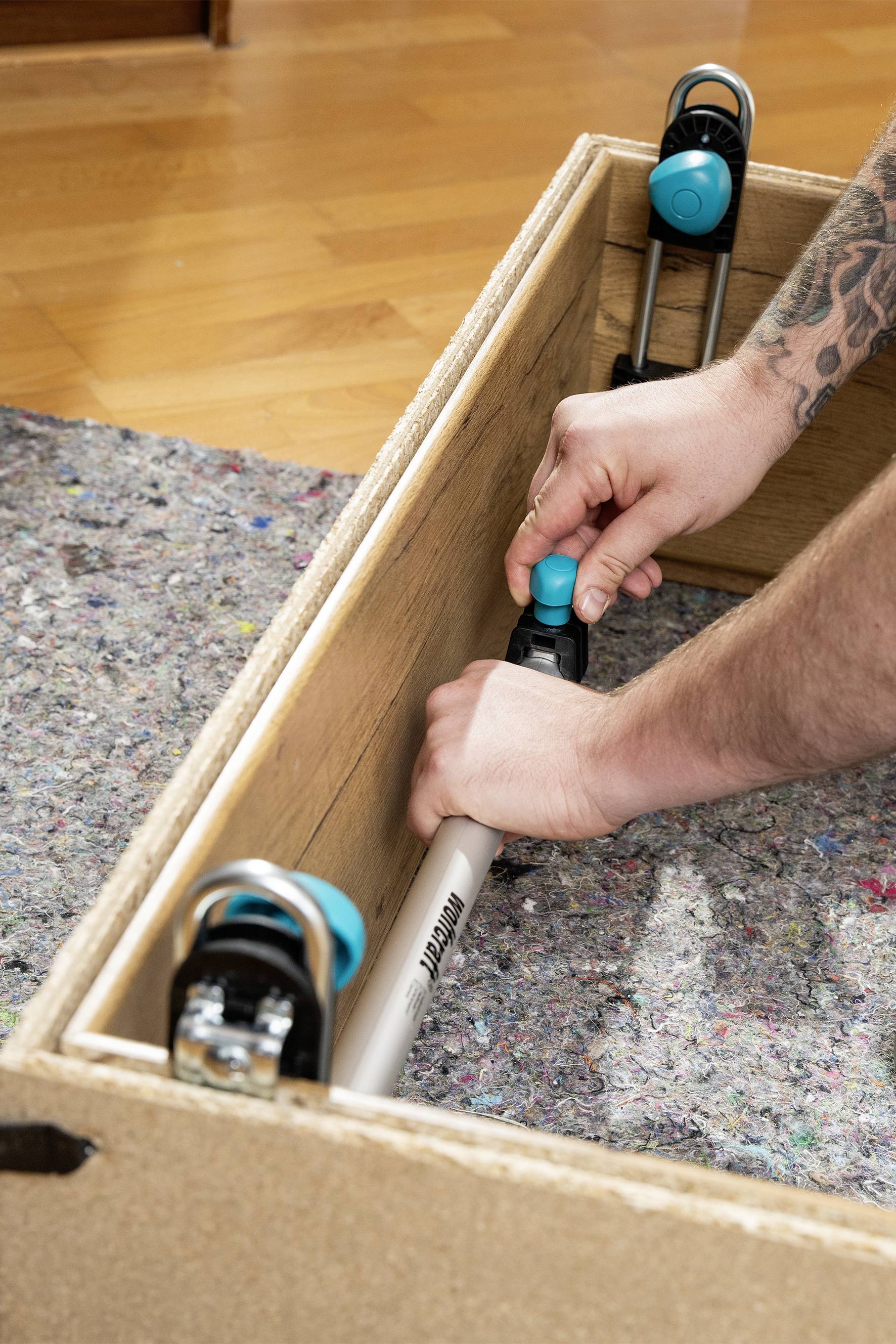 Eine Person befestigt ein Spannwerkzeug in einem Holzkasten auf einem Teppichboden. Hände ziehen das Werkzeug fest.