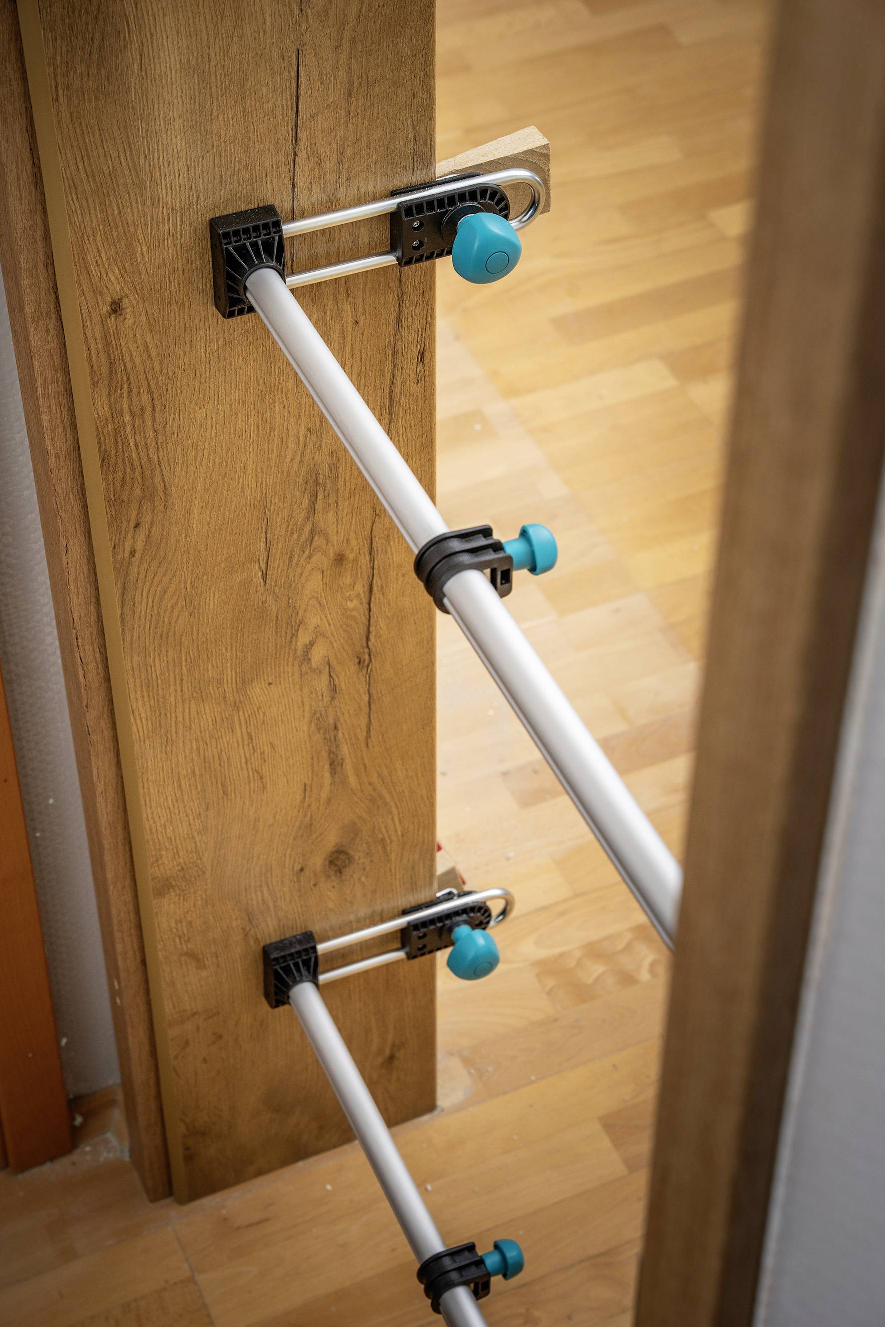 Zwei silberne Stangen sind mit blauen Klemmen an einem Holzbalken befestigt, der vor einer Holztür steht.