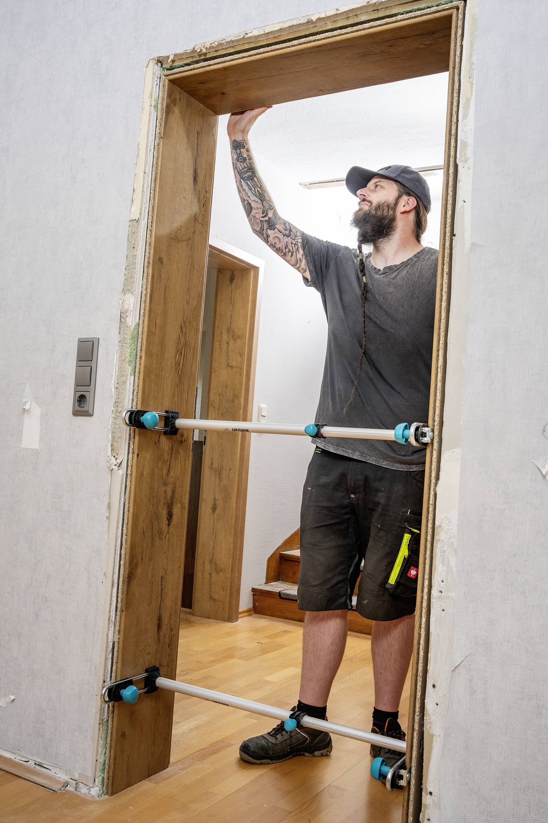 Ein Mann montiert einen Türrahmen aus Holz in einem Haus. Er trägt Arbeitskleidung und Mütze und verwendet Montagehilfen.