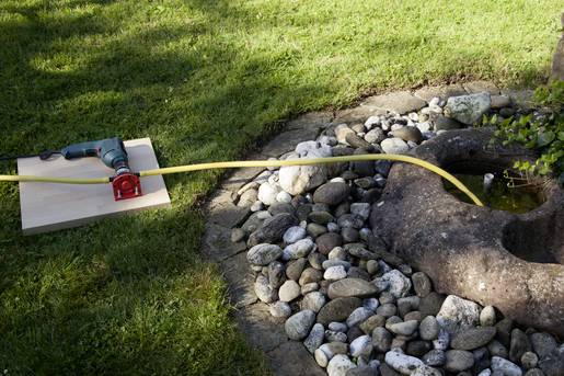 Bohrmaschine mit Schlauch pumpt Wasser in einen Steintrog im Garten, umgeben von Kieselsteinen und Rasen.