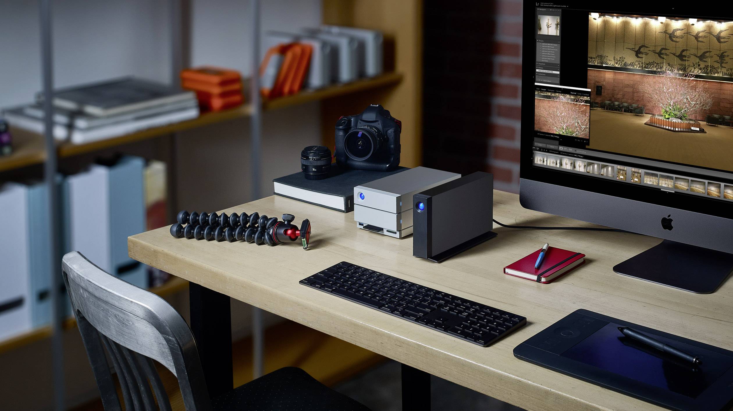 Ein Schreibtisch mit Computer und Kamera. Daneben stehen externe Festplatten und ein Stativ. Im Hintergrund ein Bücherregal.