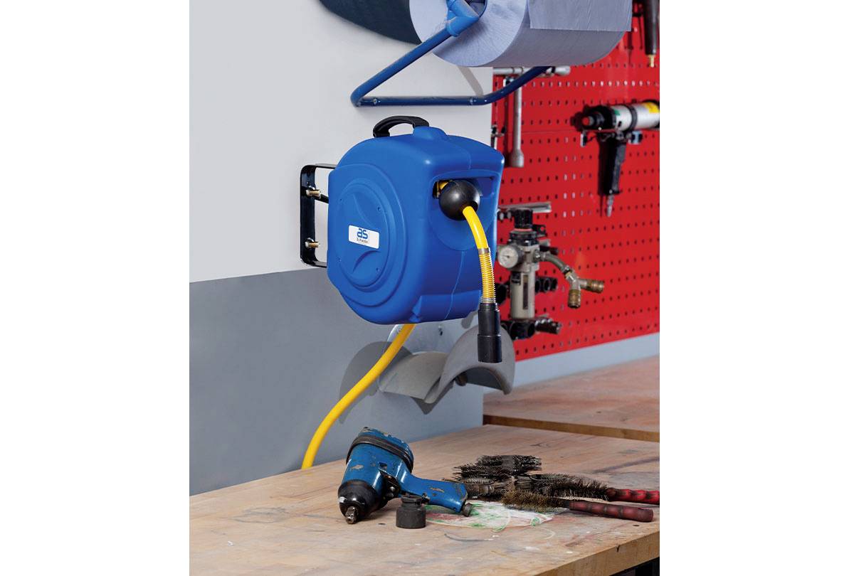Eine automatische einziehbare Druckluftschlauchrolle, an einer Werkstattwand montiert, mit einem gelben Schlauch, der mit einem blauen Druckluftgerät auf einer Holzbank verbunden ist.