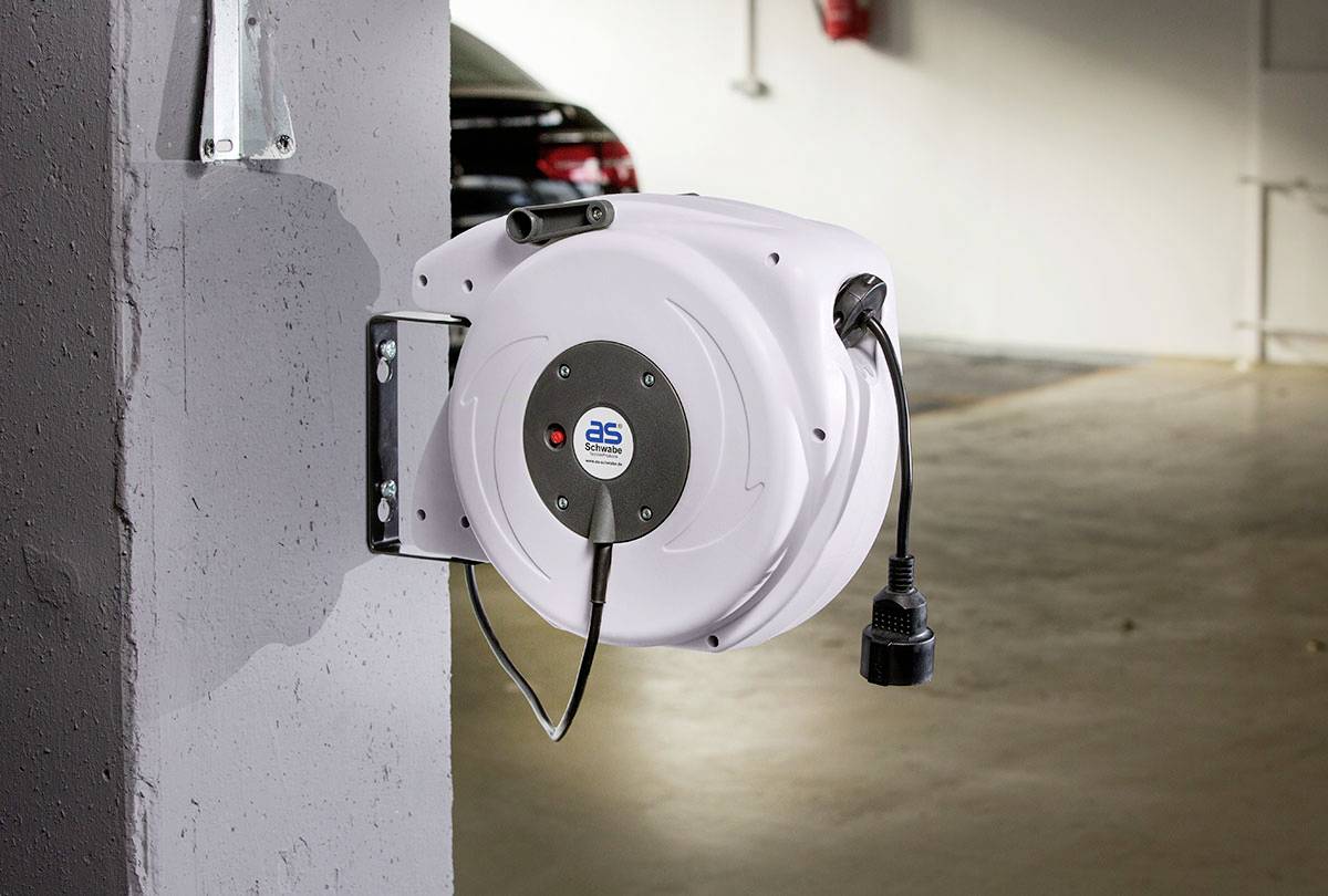 Wandmontierte Kabeltrommel in einer Tiefgarage, befestigt an einer Betonsäule; ein schwarzes Auto ist im Hintergrund zu sehen.