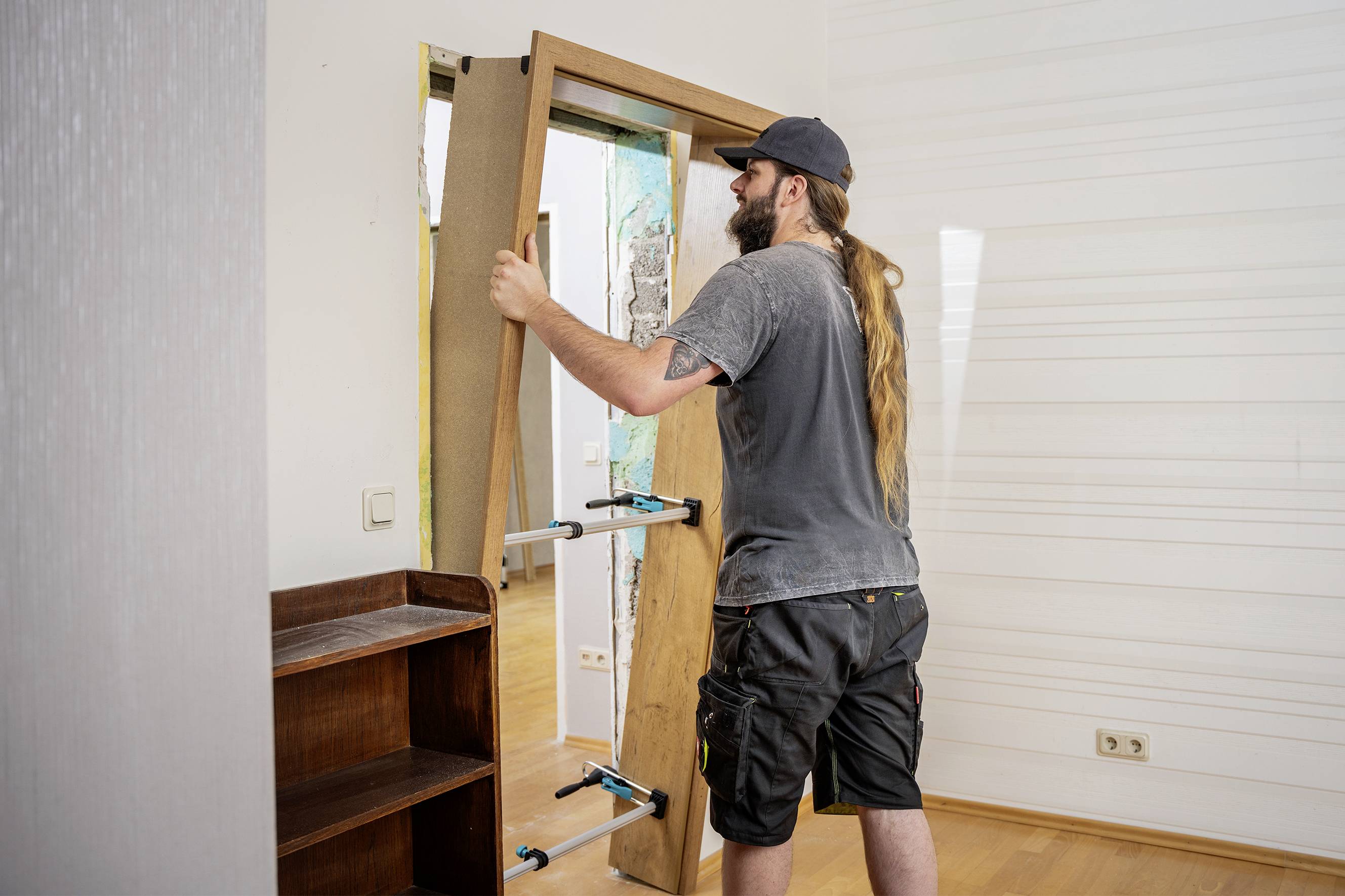 Ein Mann mit langen Haaren installiert eine Tür in einem Innenraum. Er trägt Arbeitskleidung und eine Mütze.