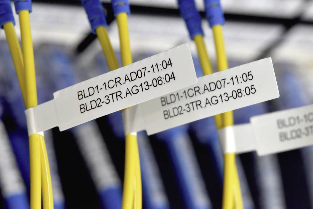 Nahaufnahme von gelben Glasfaserkabeln mit Etiketten. Etiketten zeigen numerische Codes, die auf Verbindungsdetails hinweisen. Kabel sind in einem Serverraum platziert.