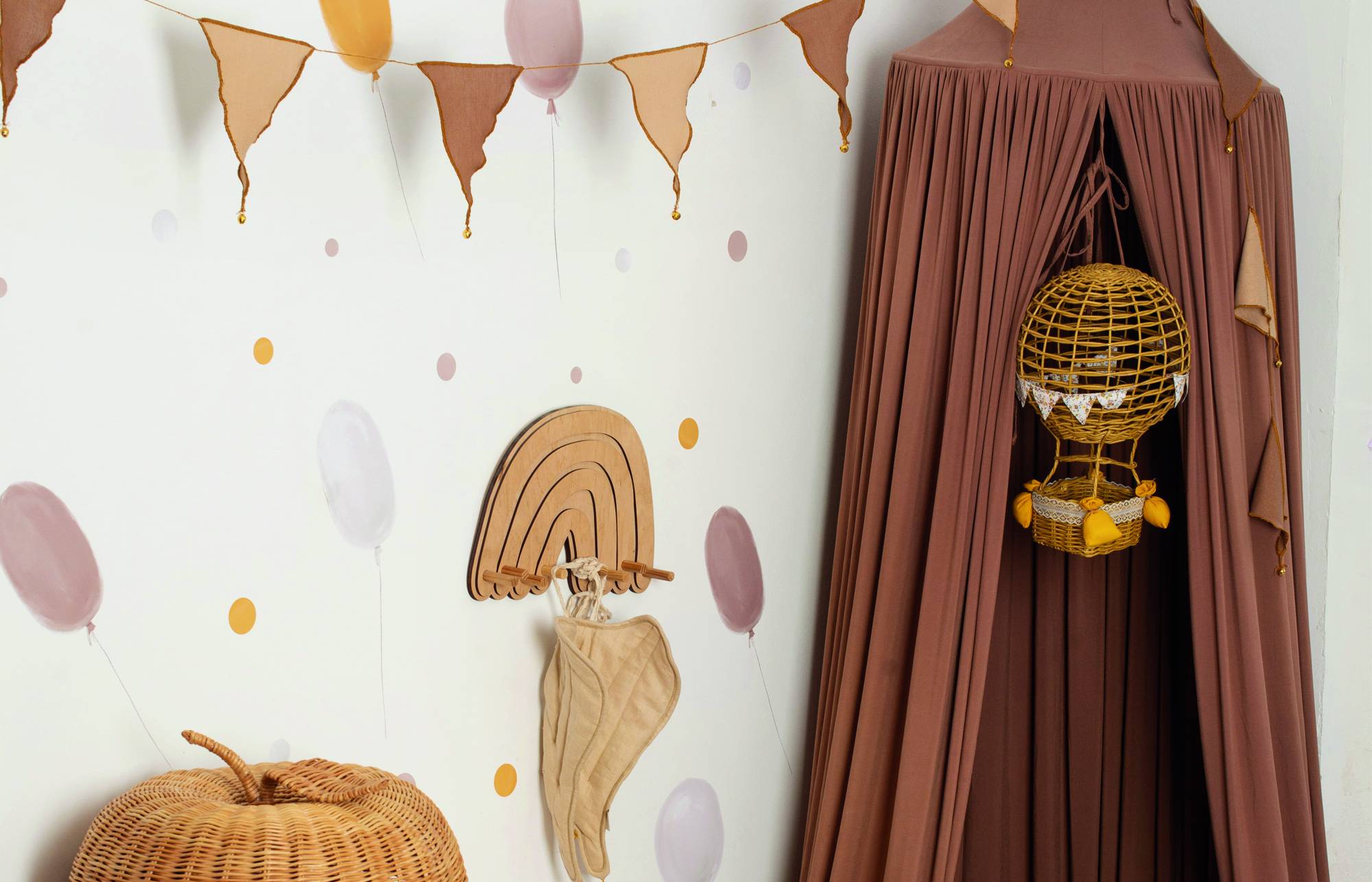 Kinderzimmerecke mit braunem Baldachin und hängendem Weiden-Heißluftballon-Spielzeug neben einer Wand, die mit Pastellballons, Wimpeln und einem Regenbogenhaken dekoriert ist.