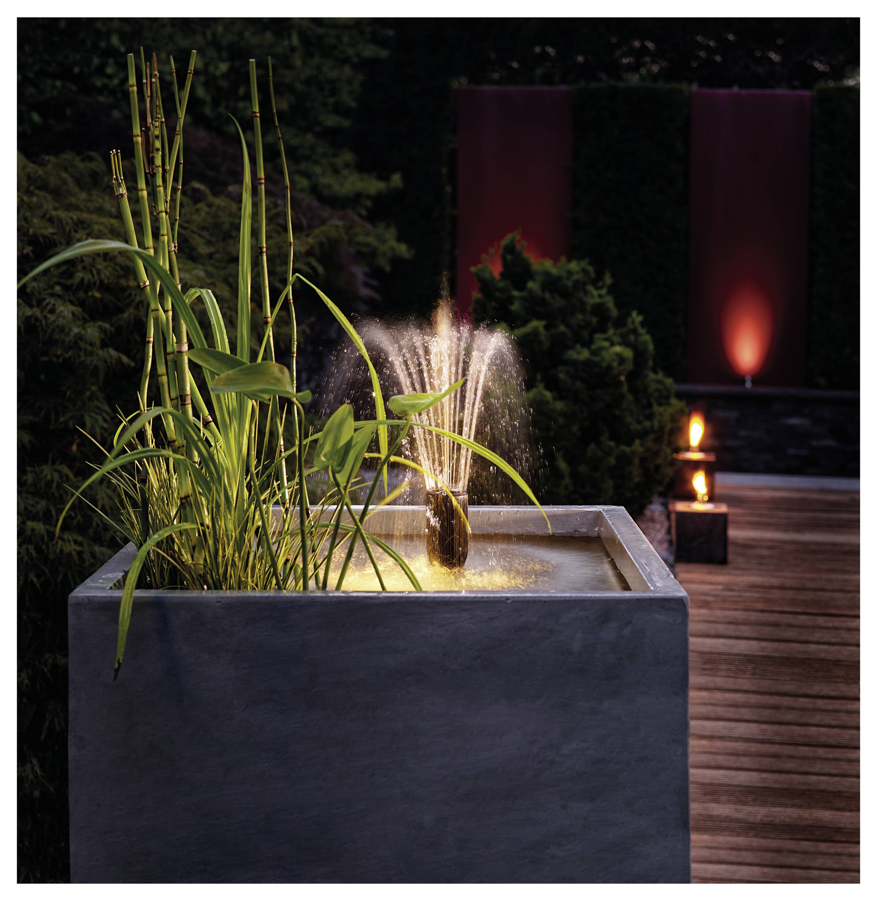 Ein moderner Brunnen mit hohen Gräsern in einem quadratischen Pflanzgefäß, der auf einer Holzterrasse mit warmem Ambientlicht in einem Garten bei Einbruch der Dämmerung steht.