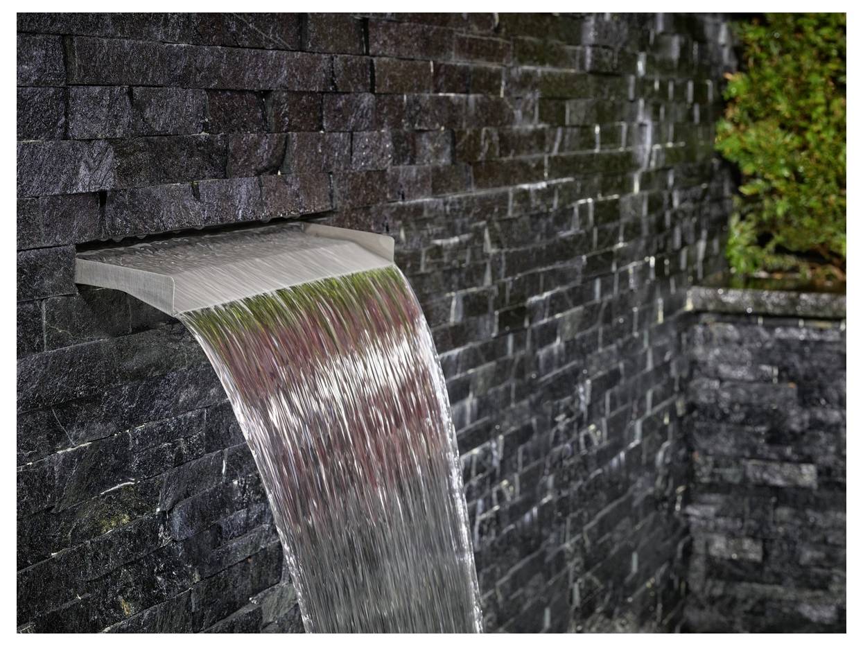 Ein sanfter Wasserstrom fließt aus einem rechteckigen Ausguss in einer Steinmauer und erzeugt einen kaskadierenden Effekt vor den dunklen strukturierten Ziegelsteinen.