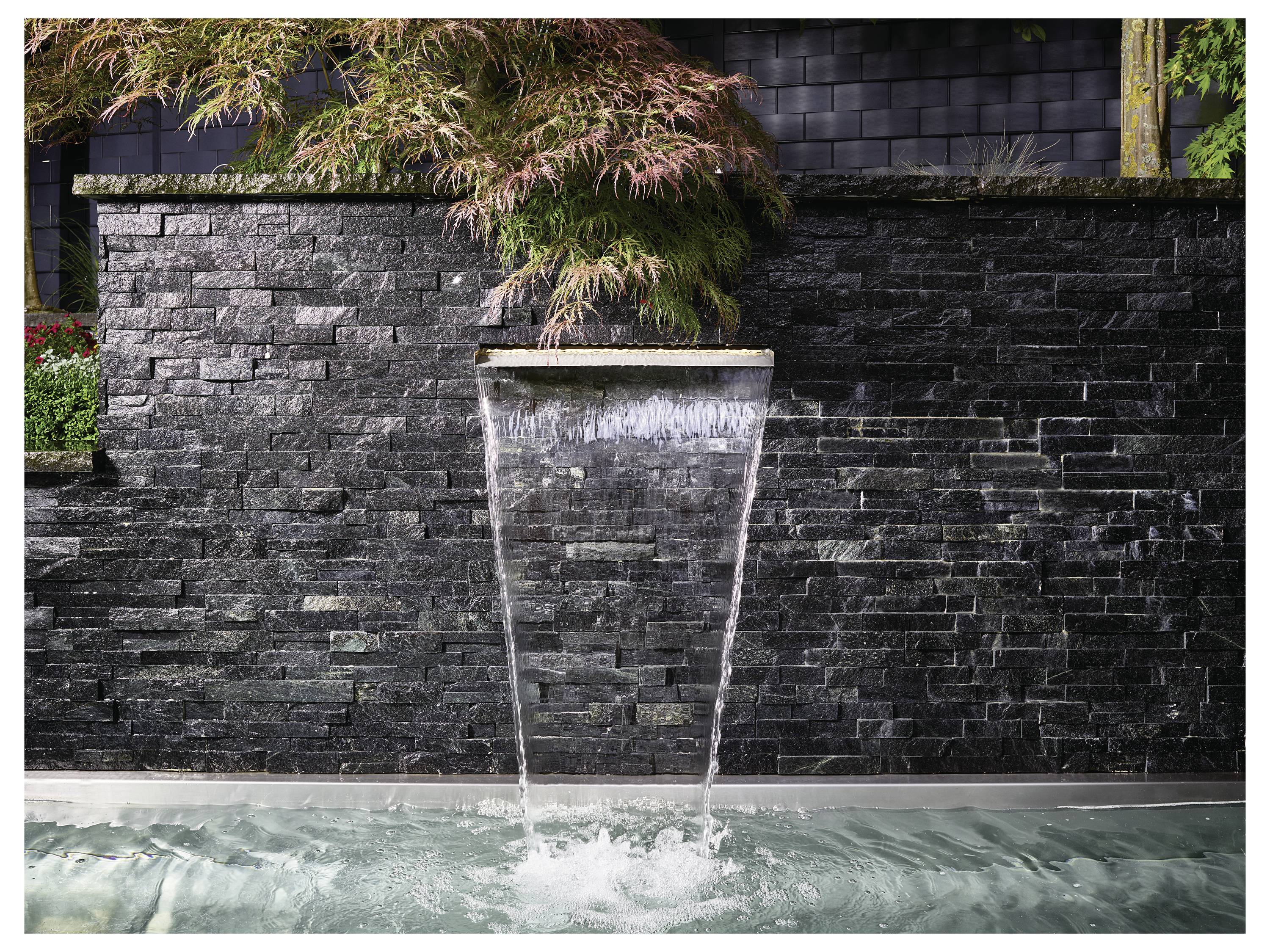 Ein modernes Wasserfallmerkmal fließt von einer Steinmauer in einen ruhigen Teich, umgeben von üppiger Vegetation und schafft eine friedvolle Atmosphäre.