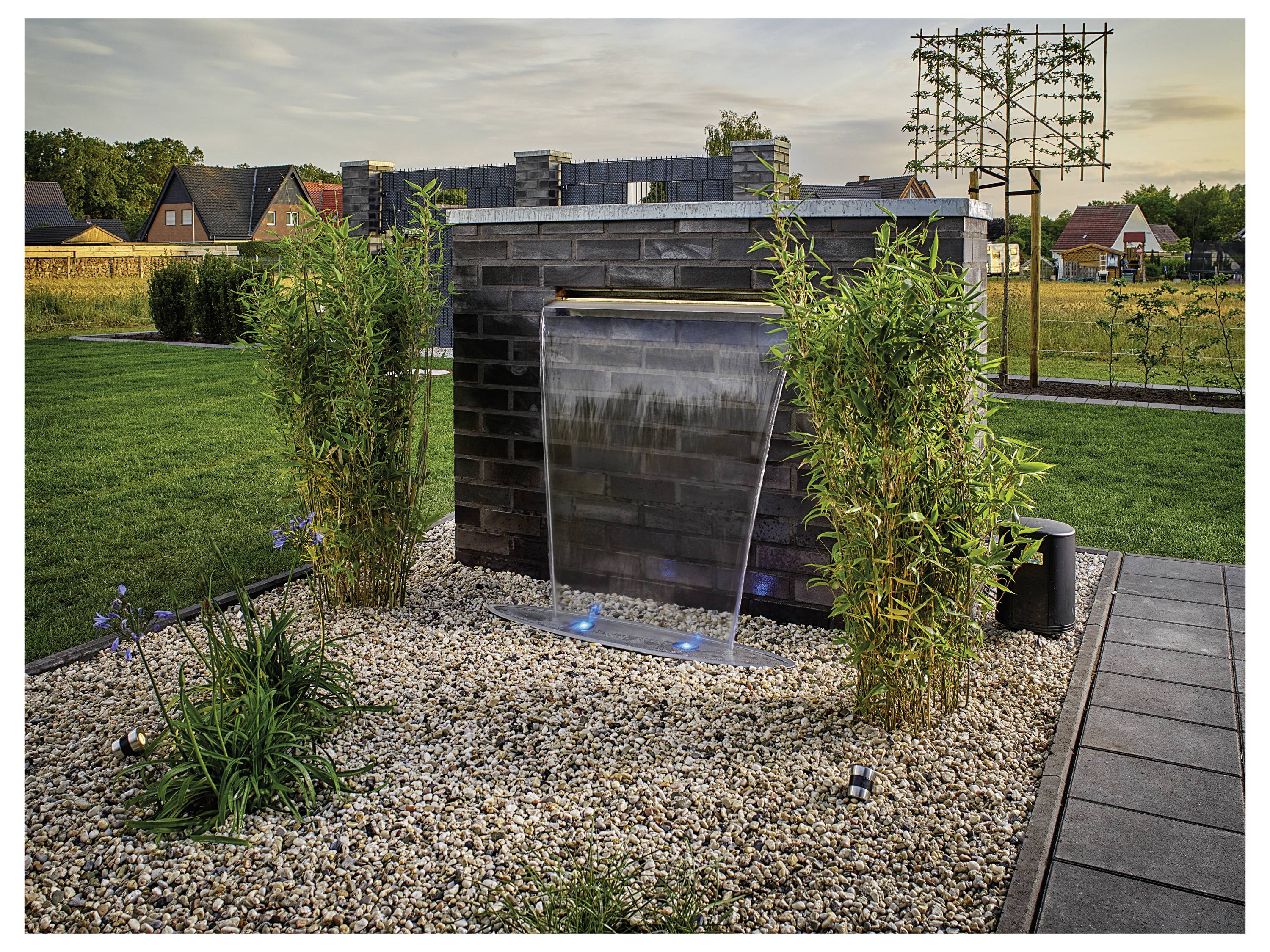 Ein kleiner Gartenwasserfall mit herabfließendem Wasser, umgeben von grünen Pflanzen und Kieselsteinen. Häuser und Bäume sind im Hintergrund zu sehen.