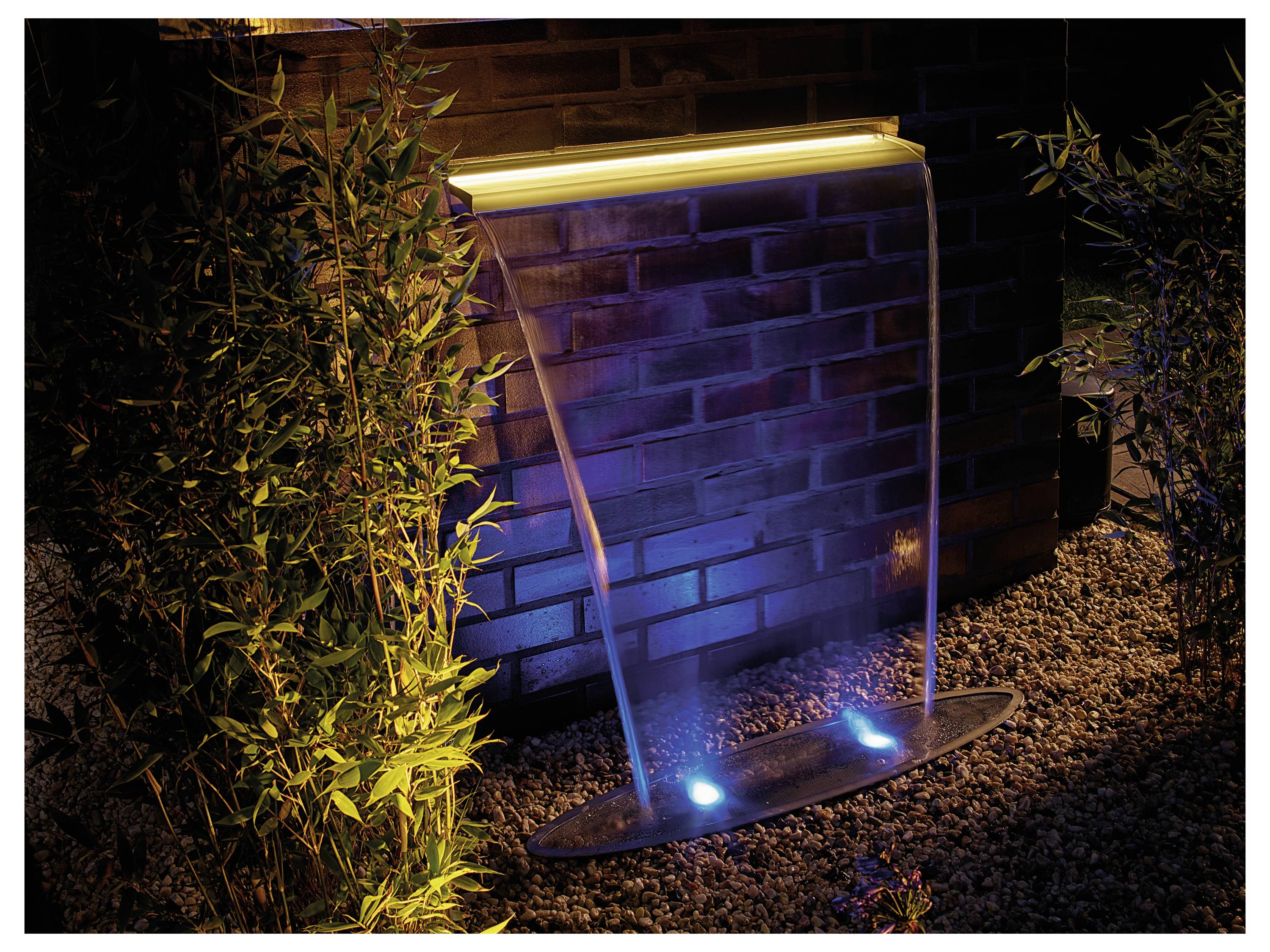 Nächtliche Szene eines modernen Brunnens vor einer Ziegelwand, mit beleuchteten Wasserstrahlen und blauen Lichtern auf einem Kiesbett.