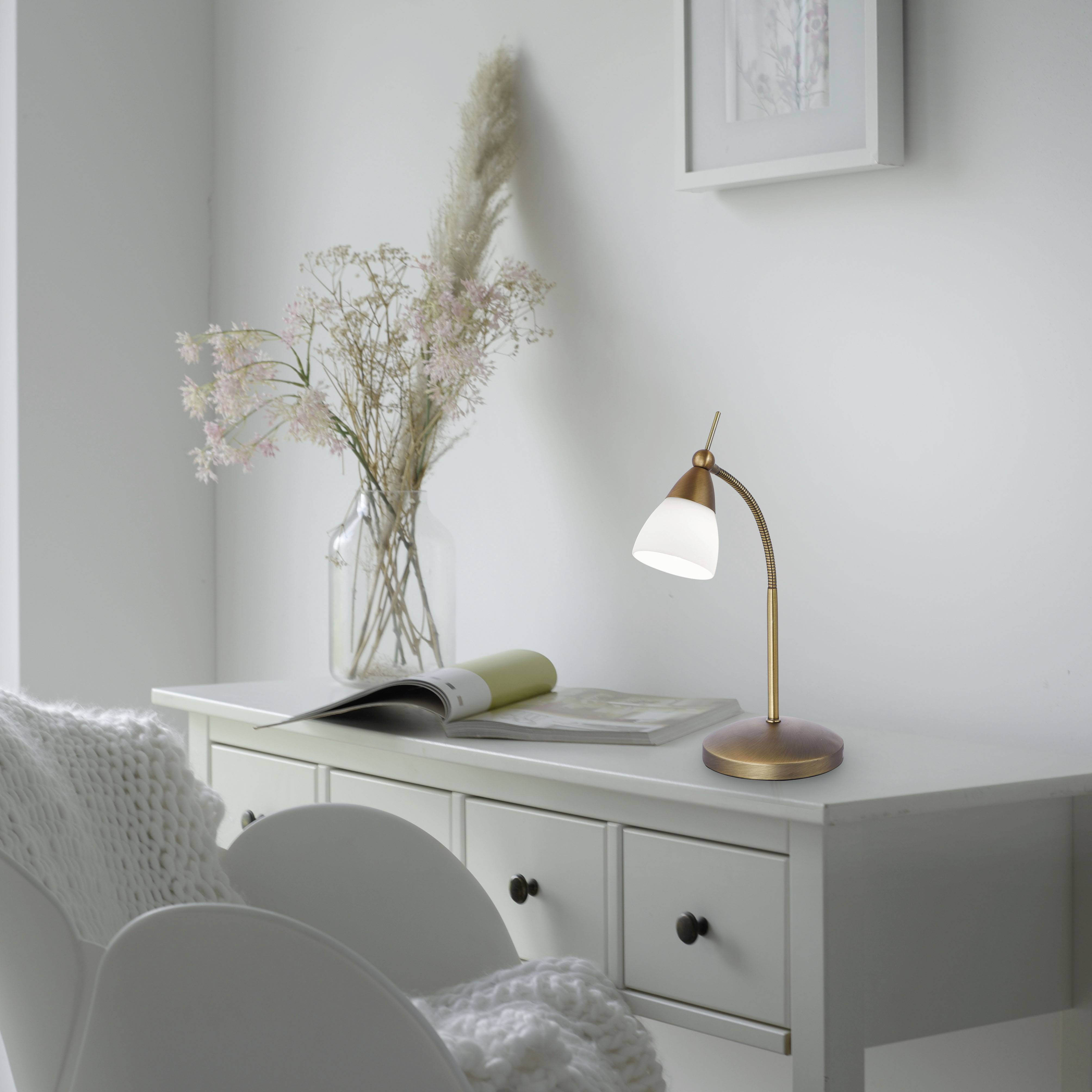 Ein heller Schreibtisch mit einer Lampe, Blumen in einer Vase und einem Stuhl mit Strickdecke, vermittelt eine entspannte Atmosphäre.