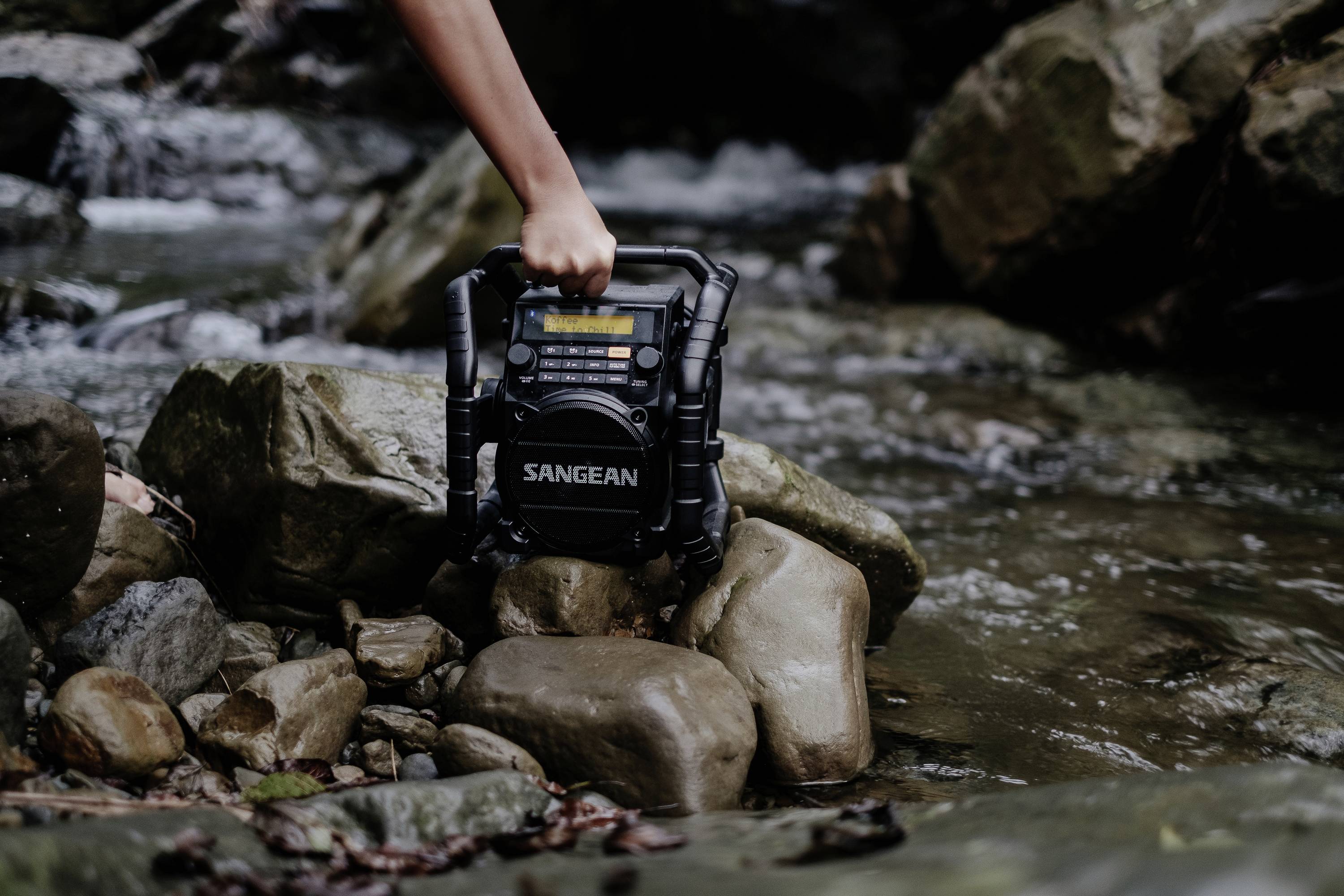 Eine Hand hält ein robustes Radio mit dem Markennamen 'Sangean', platziert auf Felsen in einem flachen Bach in einer natürlichen Umgebung.