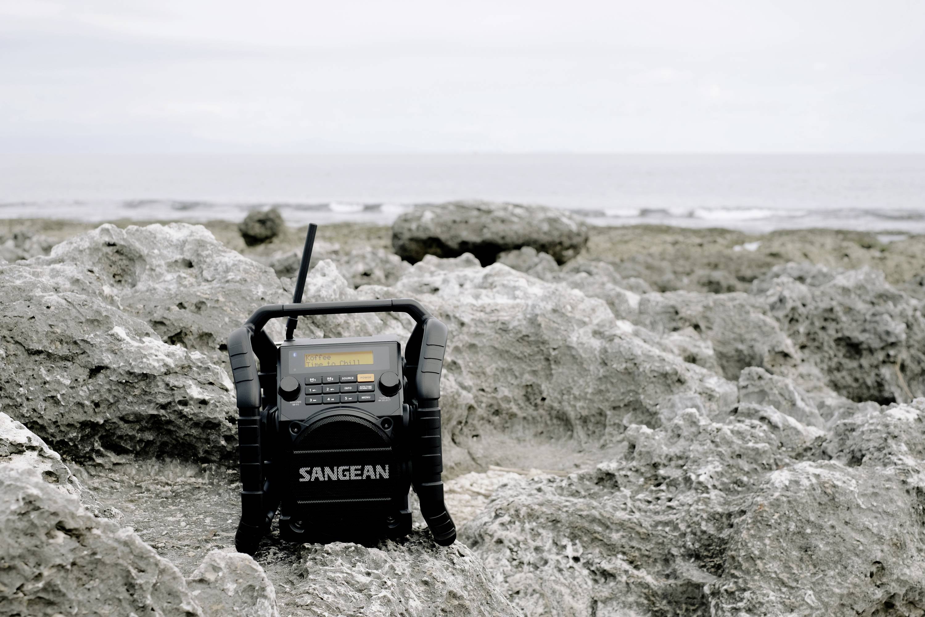 Ein tragbares Radio steht auf einer felsigen Küstenlandschaft. Der Himmel ist bewölkt, das Meer im Hintergrund ist ruhig.