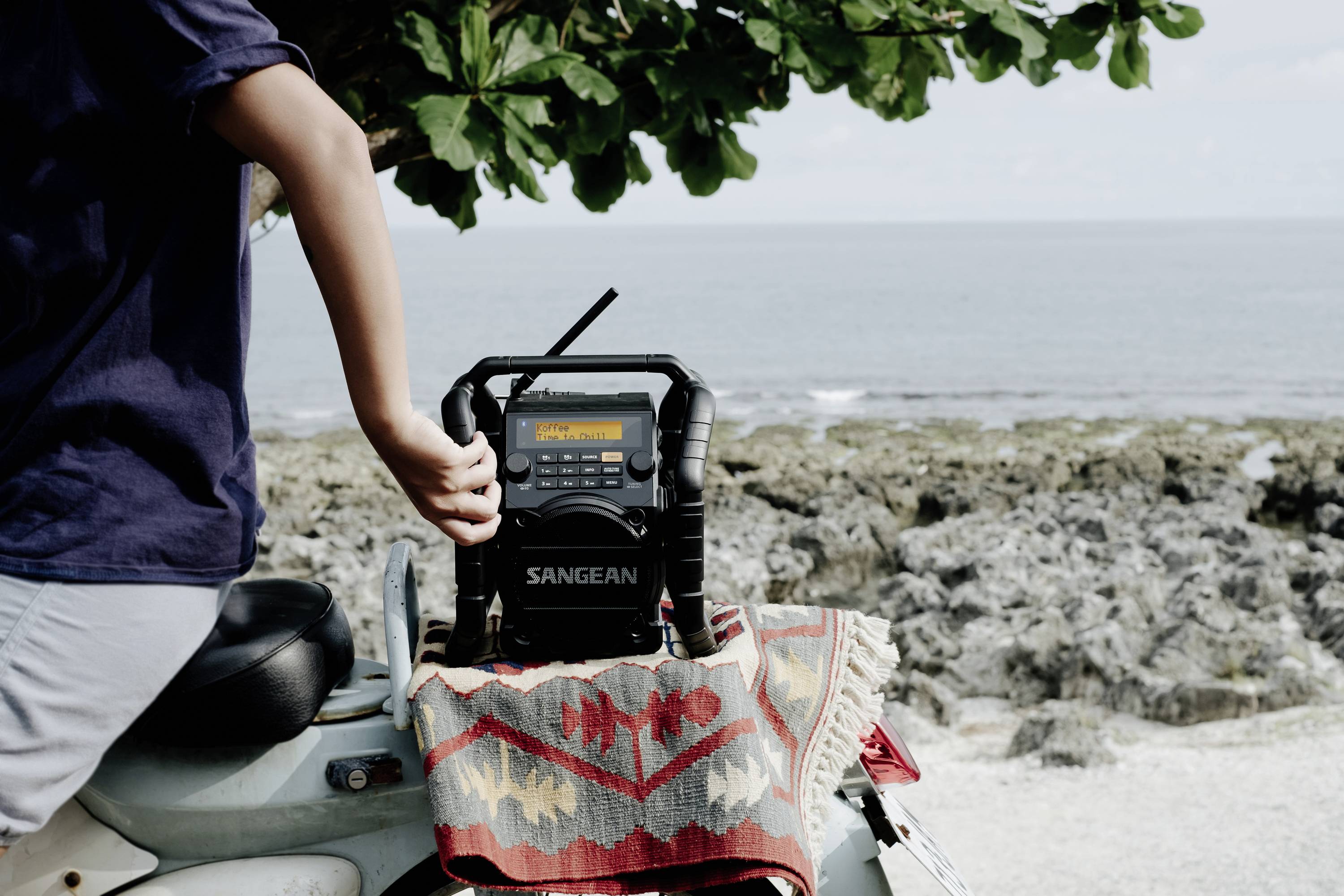 Eine Person steht neben einem Küstenabschnitt und berührt ein tragbares Radio auf einem Scooter. Meer und Felsen im Hintergrund.