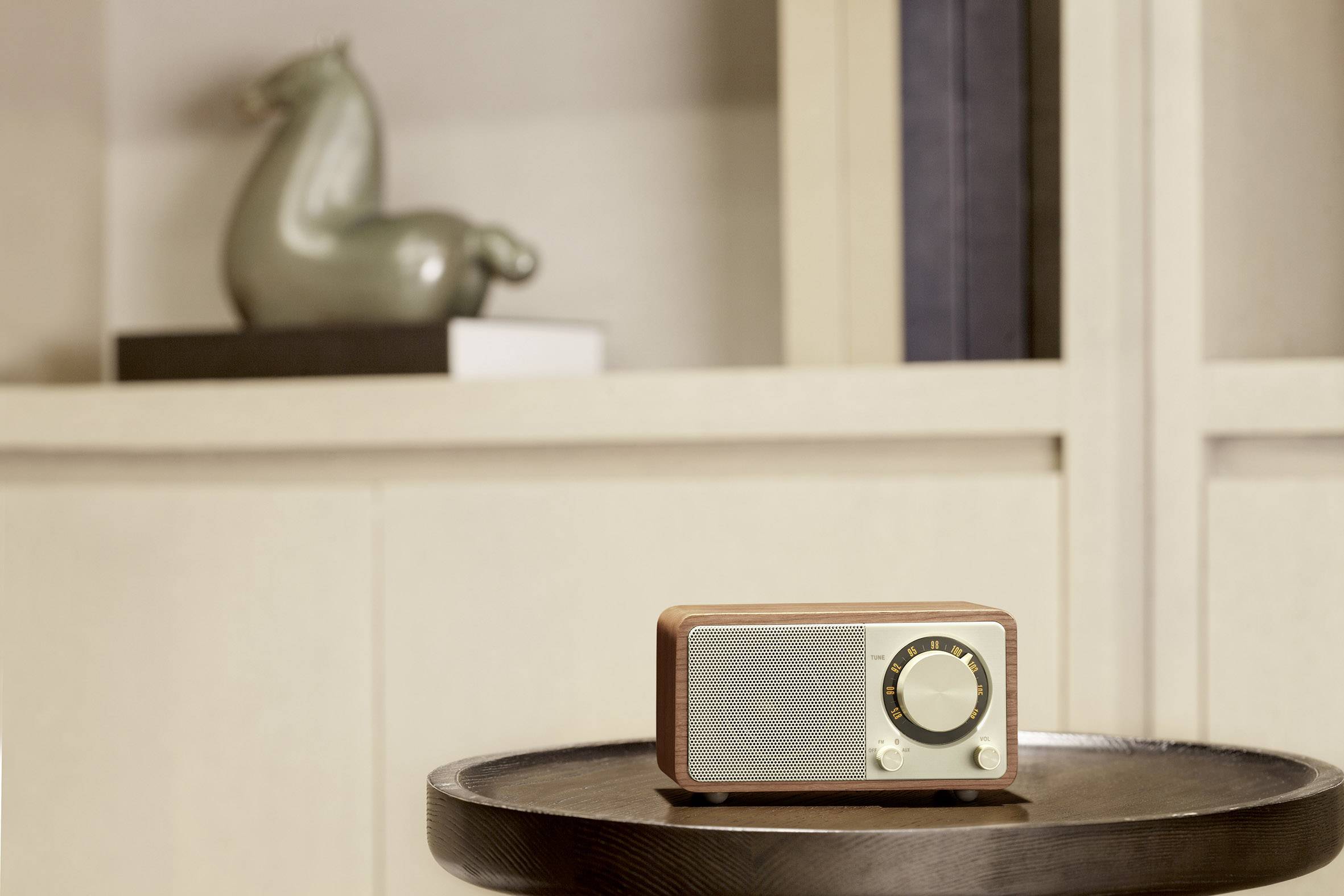 Ein kleiner Retro-Radio auf einem Holztisch vor einem weißen Regal mit dekorativer Vase im Hintergrund.