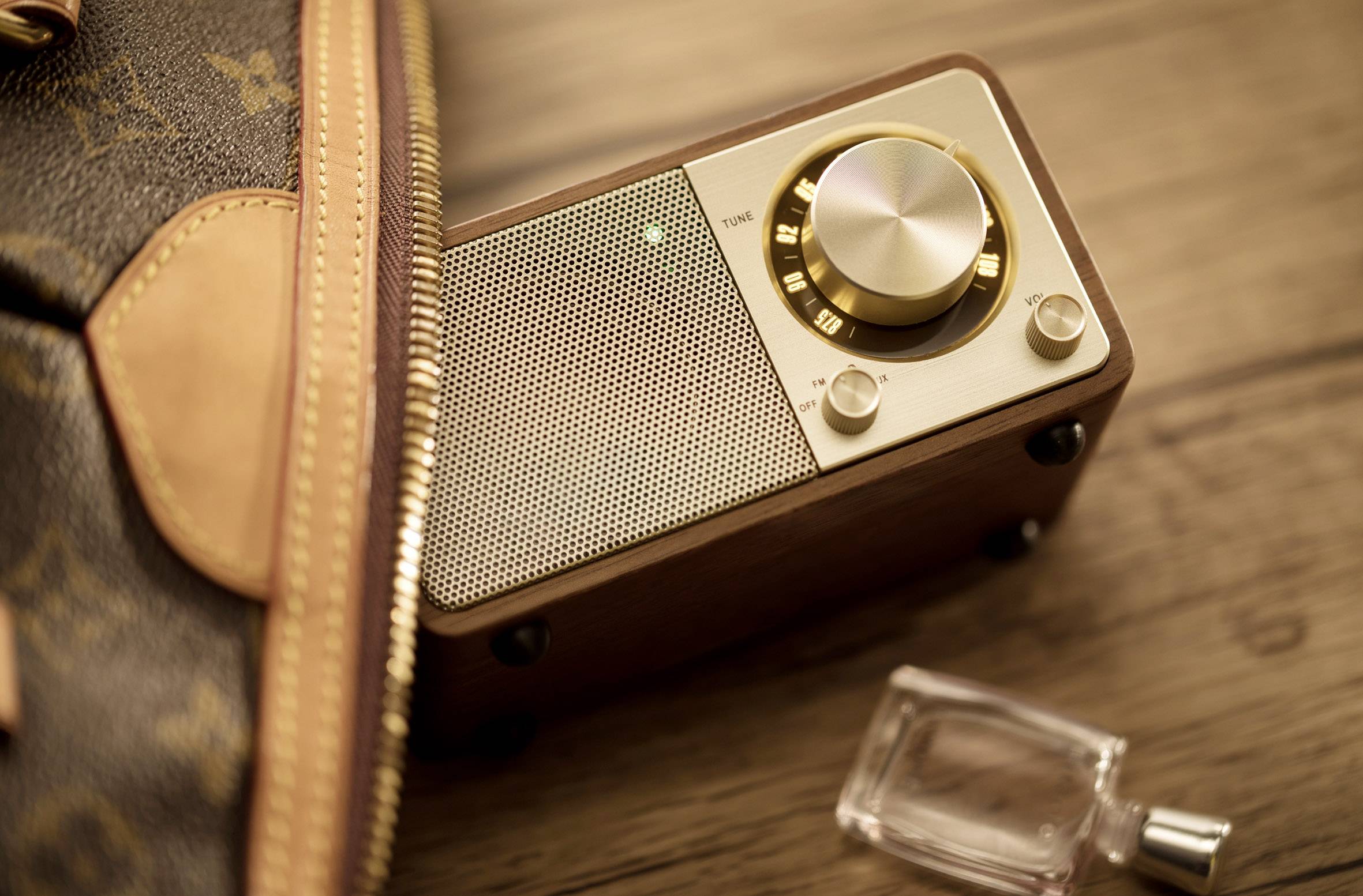 Tragbares Retro-Radio mit goldenen Knöpfen teilweise sichtbar neben einer Handtasche auf einem Holztisch, daneben eine Parfümflasche.