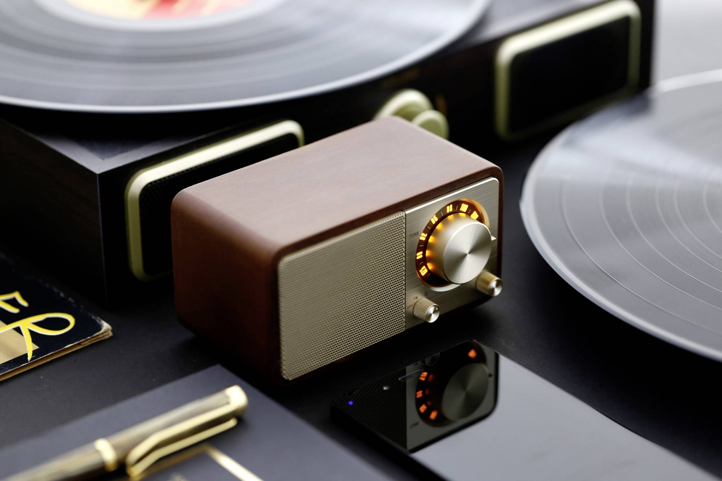 Kleiner, eleganter Lautsprecher im Retro-Design mit Holzgehäuse und beleuchtetem Lautstärkeregler, umgeben von Schallplatten.