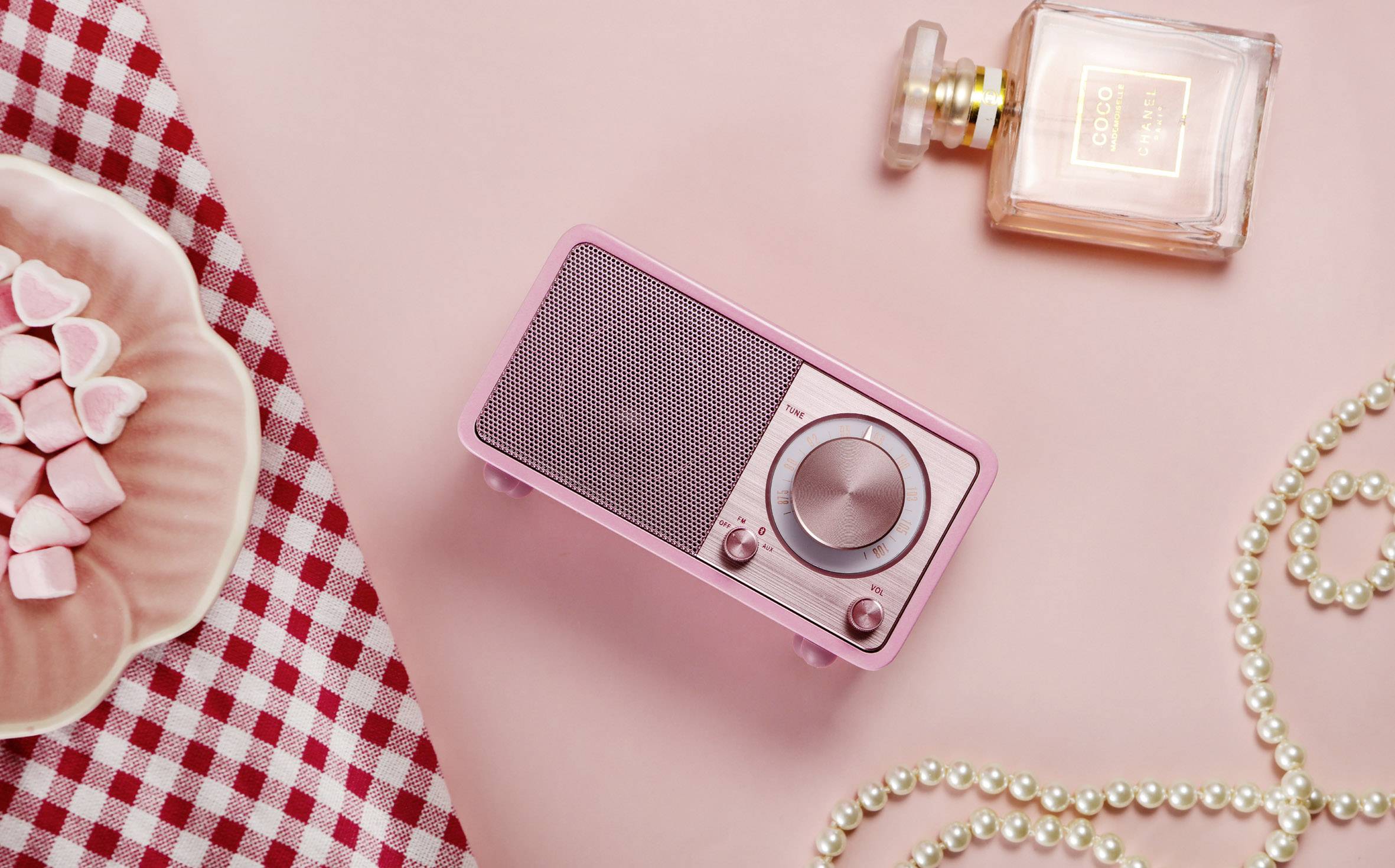 Ein rosa Retro-Radio, eine Parfümflasche, Herz-Bonbons auf einem Teller und eine Perlenkette liegen auf einem rosa Hintergrund.