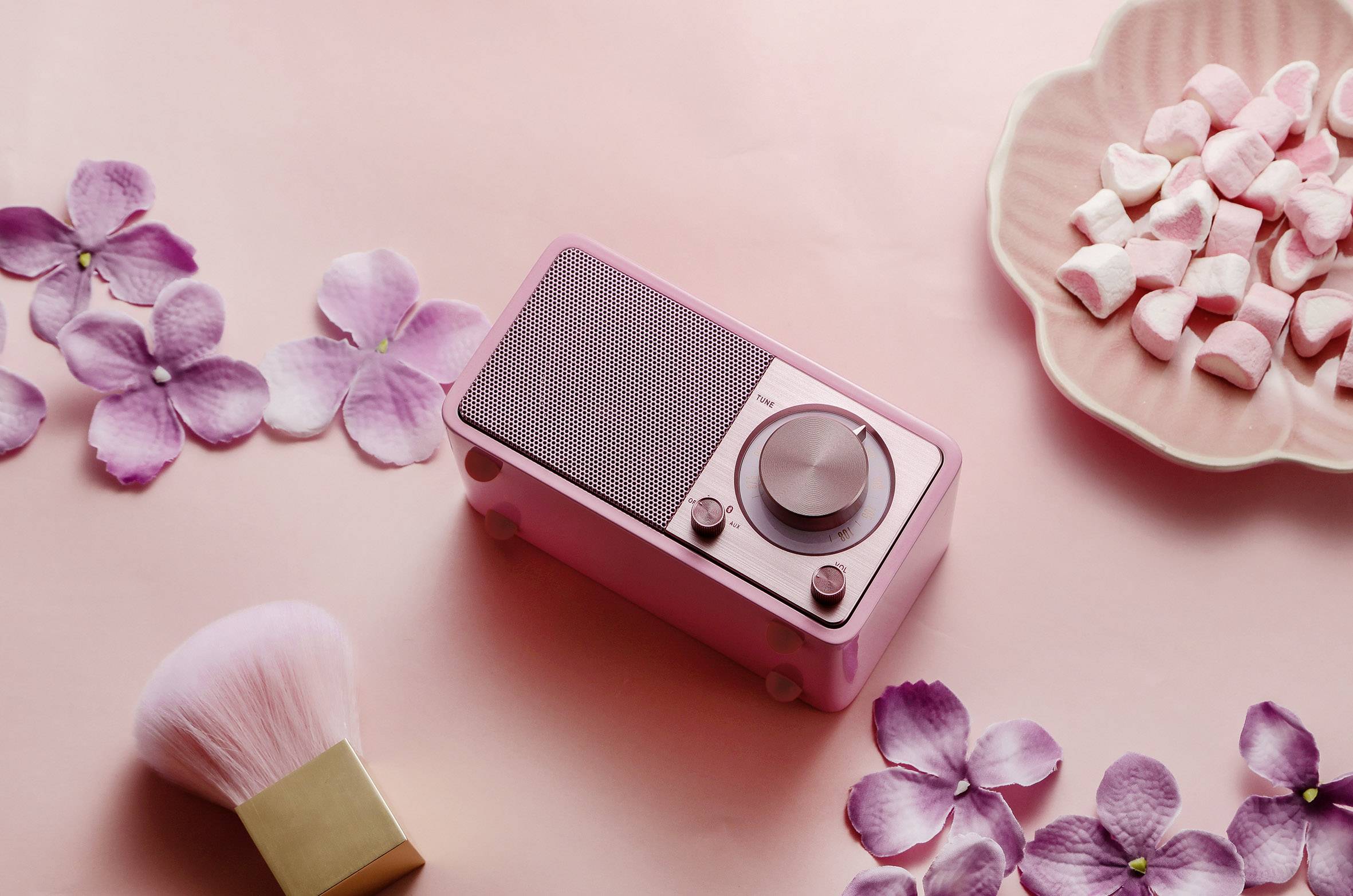 Kleines rosa Radio auf rosa Hintergrund mit Blüten und Süßigkeiten. Hauptthema: Retro-Design und Pastellfarben-Ästhetik.