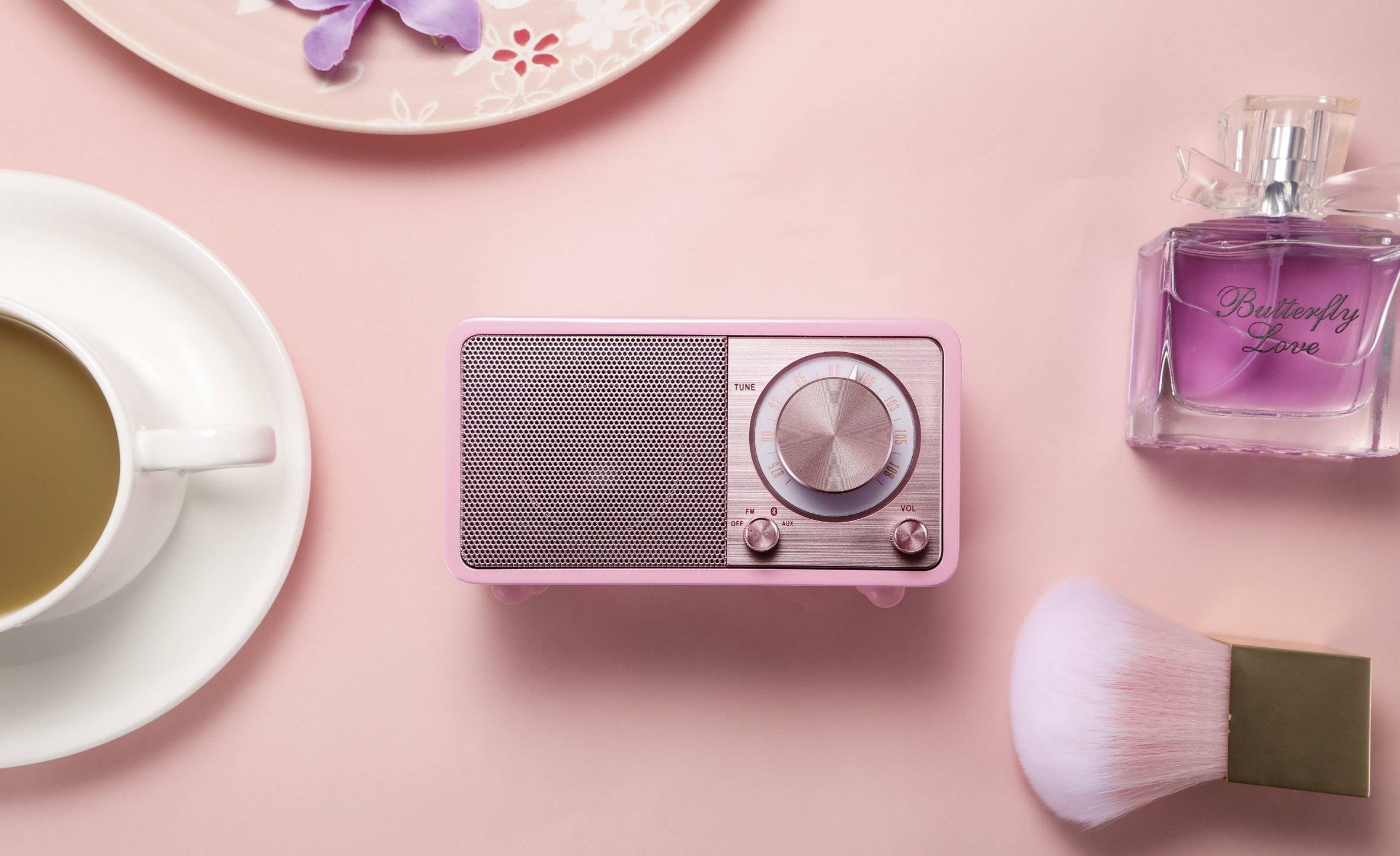 Rosa Vintage-Radio auf rosa Hintergrund, umgeben von Kaffeeschale, Parfümflasche und Make-up-Pinsel.