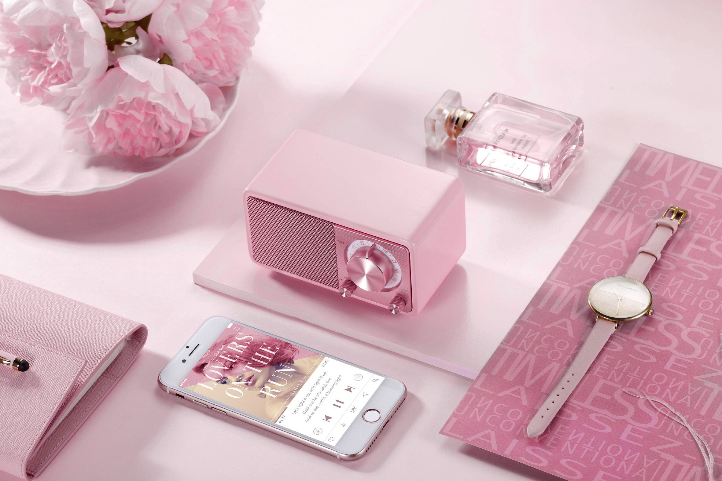 Auf einem rosa Hintergrund liegen ein kleines rosa Radio, ein Parfümflakon, ein Smartphone mit geöffneter Musik-App, eine Armbanduhr und ein Notizbuch.