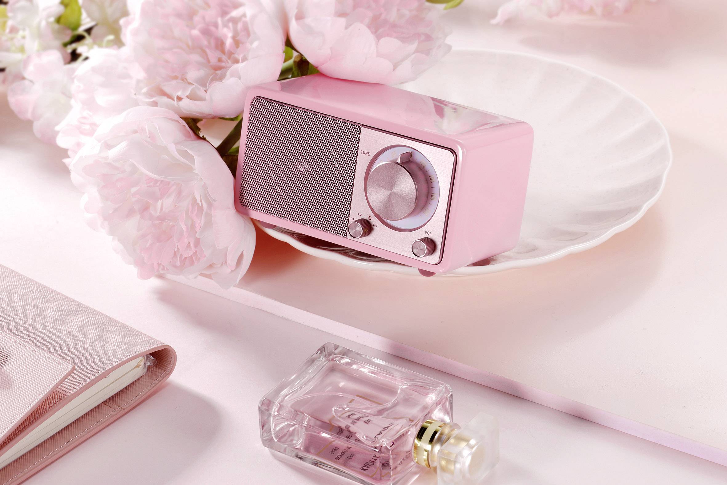 Ein rosa Retro-Radio steht auf einem Teller, umgeben von rosa Pfingstrosen. Ein Parfümflakon liegt daneben auf dem Tisch.