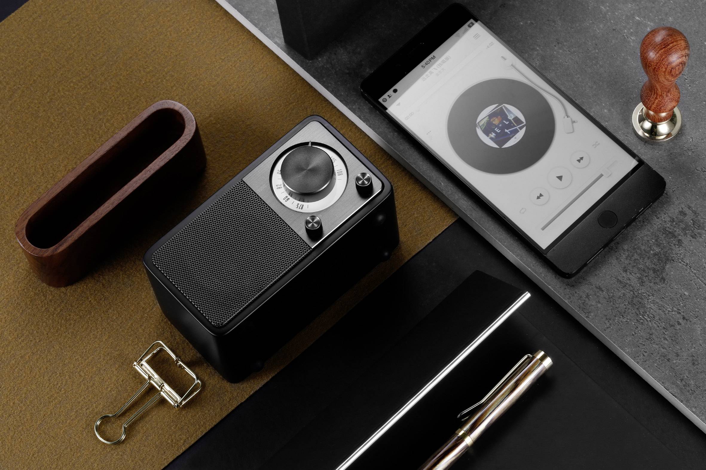 Auf einem Tisch liegen ein Lautsprecher, ein Smartphone mit Musik-App, ein Stempelsiegel, Klammern und ein Stift auf Notizblock.