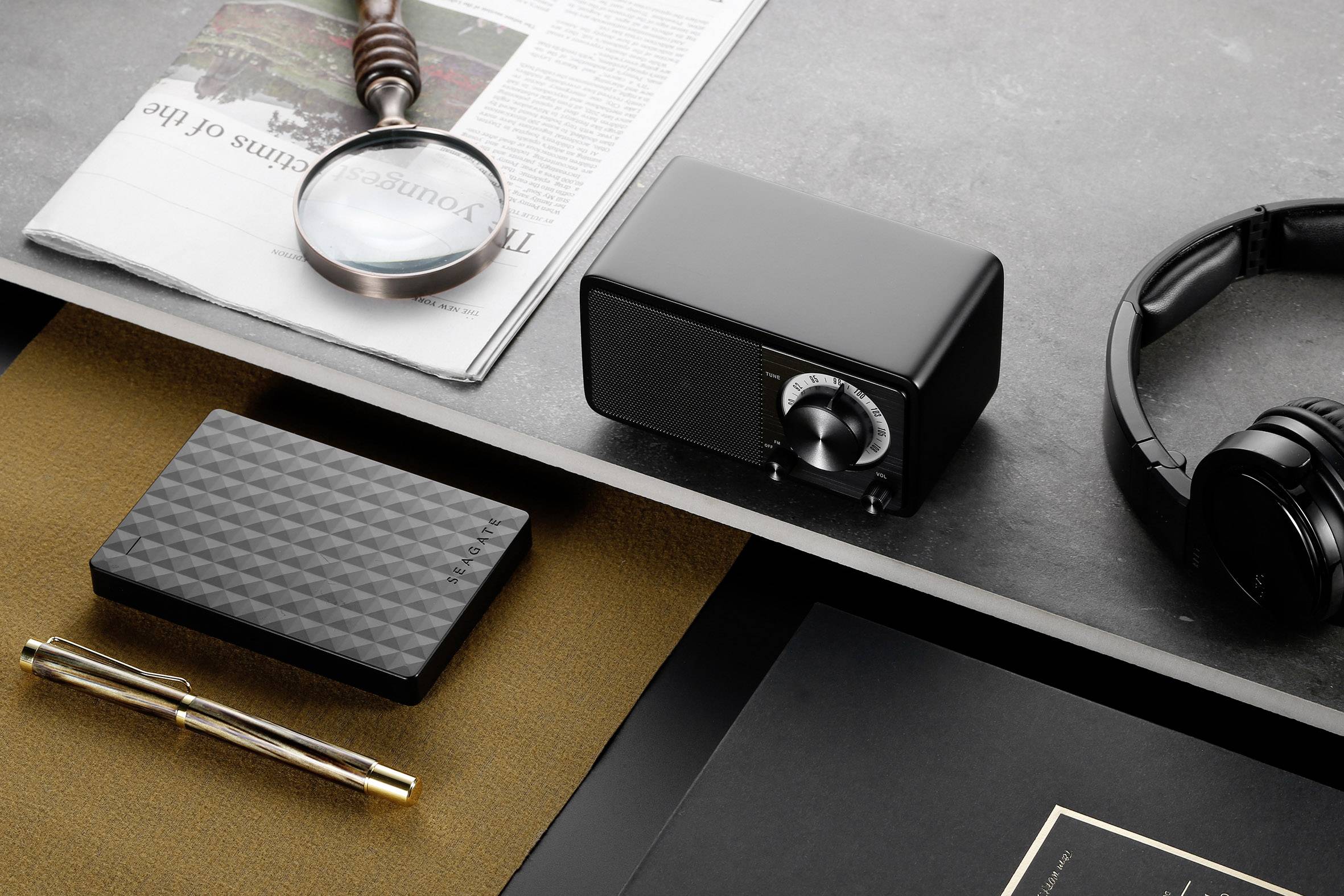 Schreibtisch mit Lupe auf Zeitung, schwarzem Radio, Kopfhörern, Stift und Festplatte, daneben ein schwarzes Notizbuch.