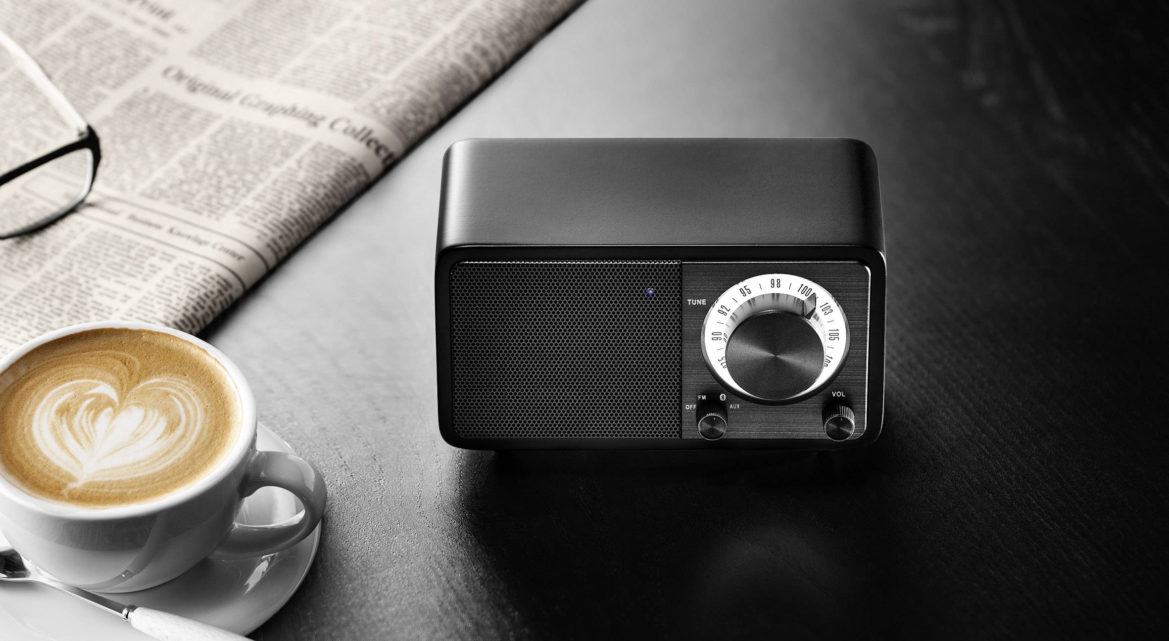 Ein schwarzes Radio mit Drehregler auf einem Tisch neben einer Tasse Cappuccino und einer Zeitung.