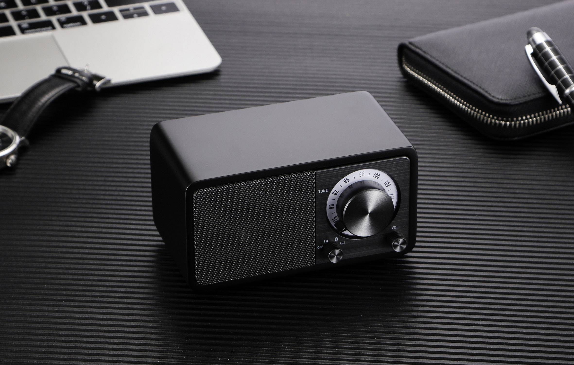 Ein schwarzer, rechteckiger Lautsprecher mit Drehknopf und Skala auf einem Tisch. Daneben ein Laptop, eine schwarze Tasche und ein Stift.