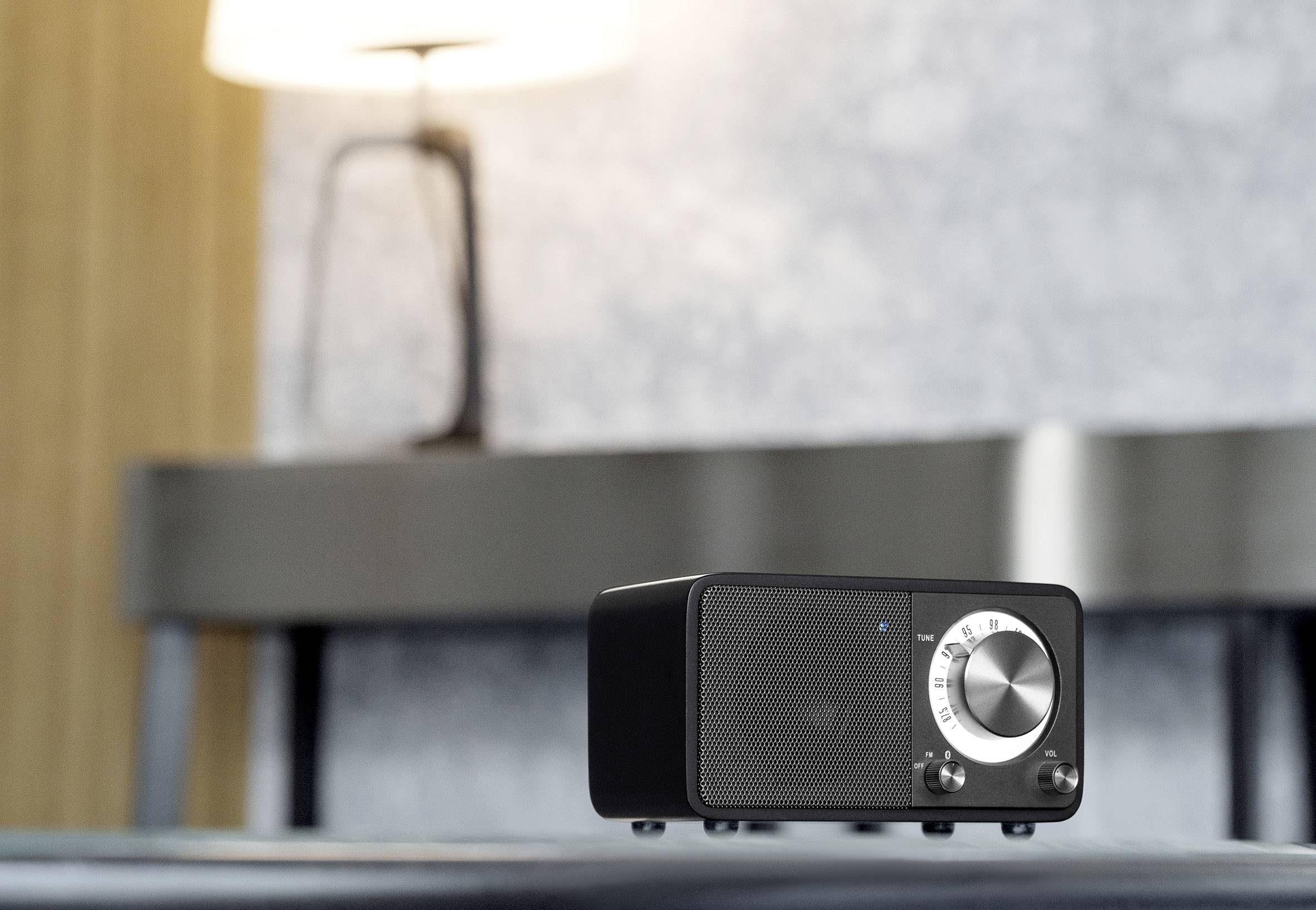 Ein moderner, schwarzer Lautsprecher mit silbernem Drehknopf steht in einem stilvoll eingerichteten Raum auf einem Tisch. Im Hintergrund ist eine Lampe unscharf zu sehen.
