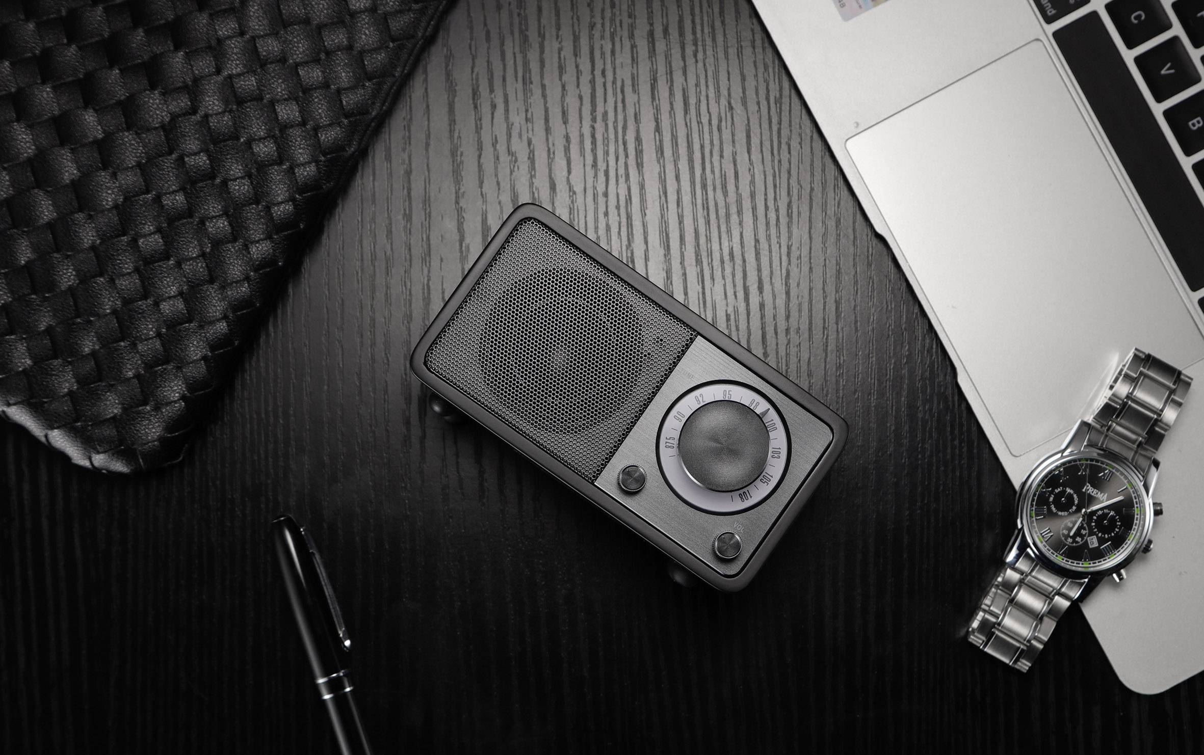 Ein schwarzes Tischradio mit Lautsprechern und Reglern sitzt auf einem dunklen Holztisch neben einem Laptop, einer Uhr und einem Stift.