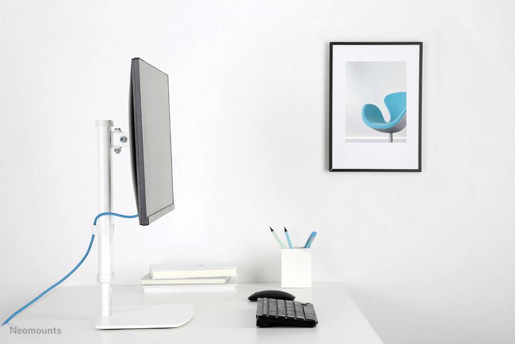 Ein moderner Arbeitsbereich mit einem seitlich montierten Monitor auf einem weißen Tisch. Daneben stehen Bücher, Stifte und eine Tastatur. An der Wand hängt ein Bild eines blauen Stuhls.