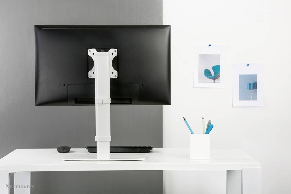 Ein Schreibtisch mit einem Monitor auf einem weißen Tisch. Daneben ein Behälter mit blauen Stiften. An der Wand hängen zwei Bilder.