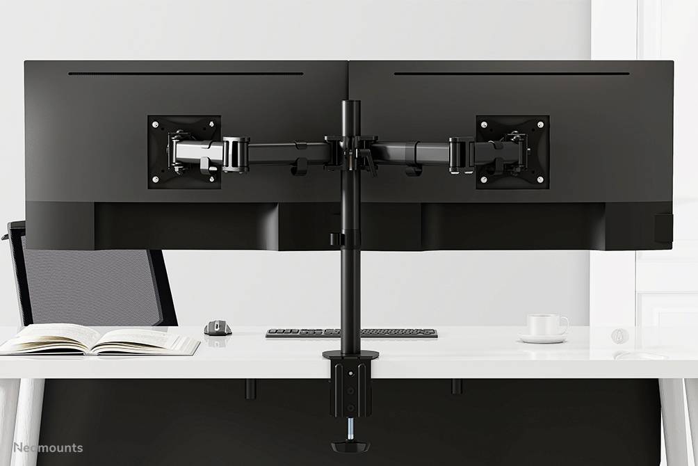 Zwei schwarze Monitore auf einer Halterung, montiert auf einem Schreibtisch. Daneben liegen ein offenes Buch, eine Maus und eine Tasse.