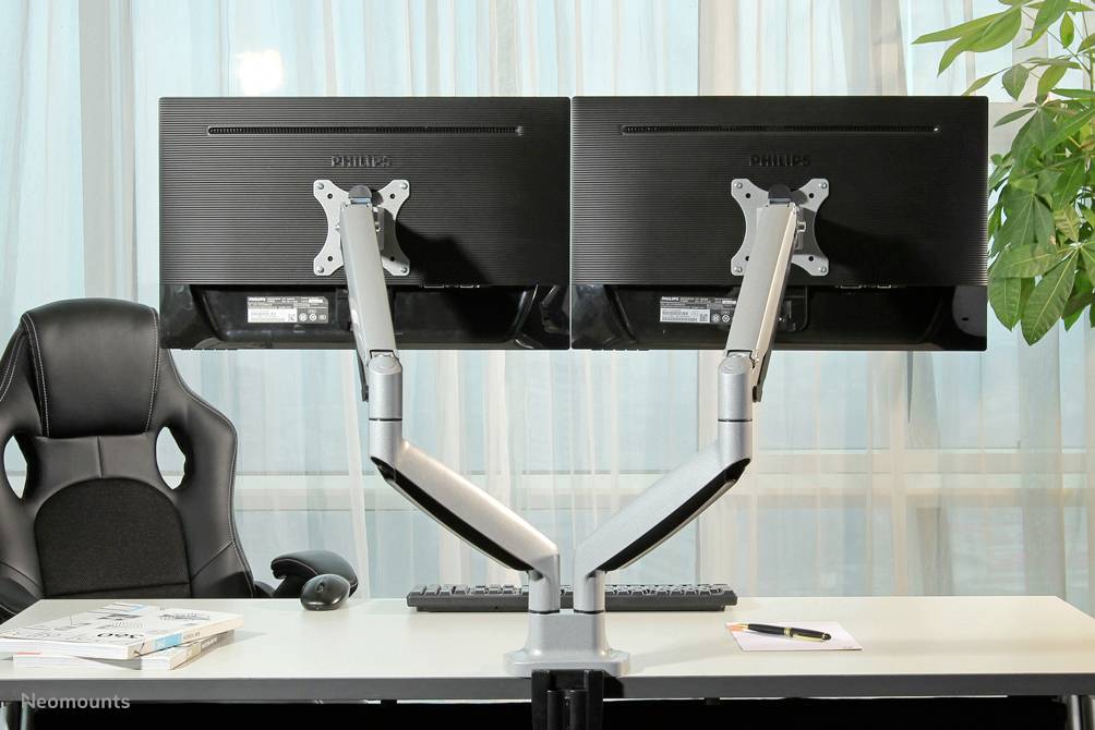 Zwei Monitore an Schwenkarmen auf einem Schreibtisch mit Bürostuhl und Pflanzen im Hintergrund. Tastatur, Maus und Notizblock vorhanden.