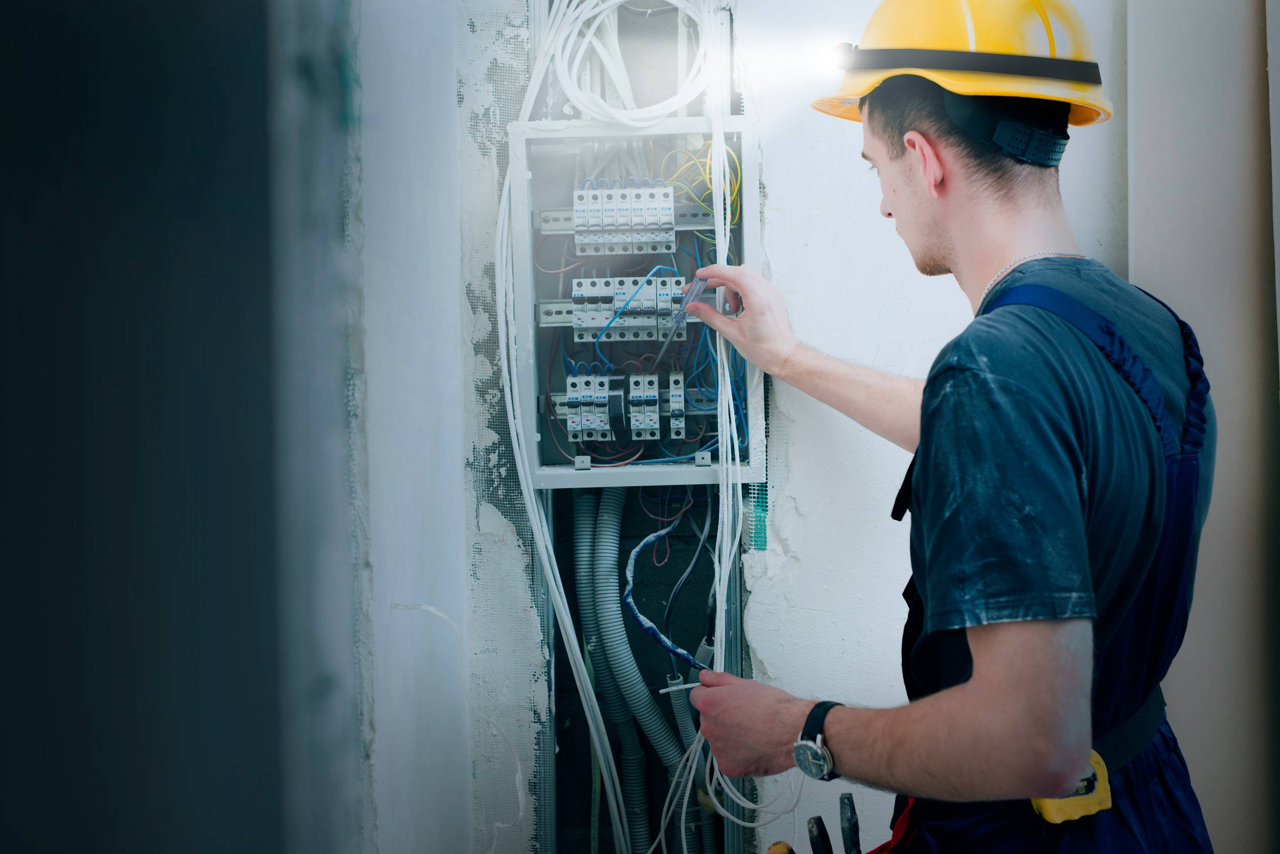 Ein Elektriker mit Helm arbeitet an einem offenen Schaltschrank, überprüft Kabelverbindungen, in einem unvollständigen Wandabschnitt.