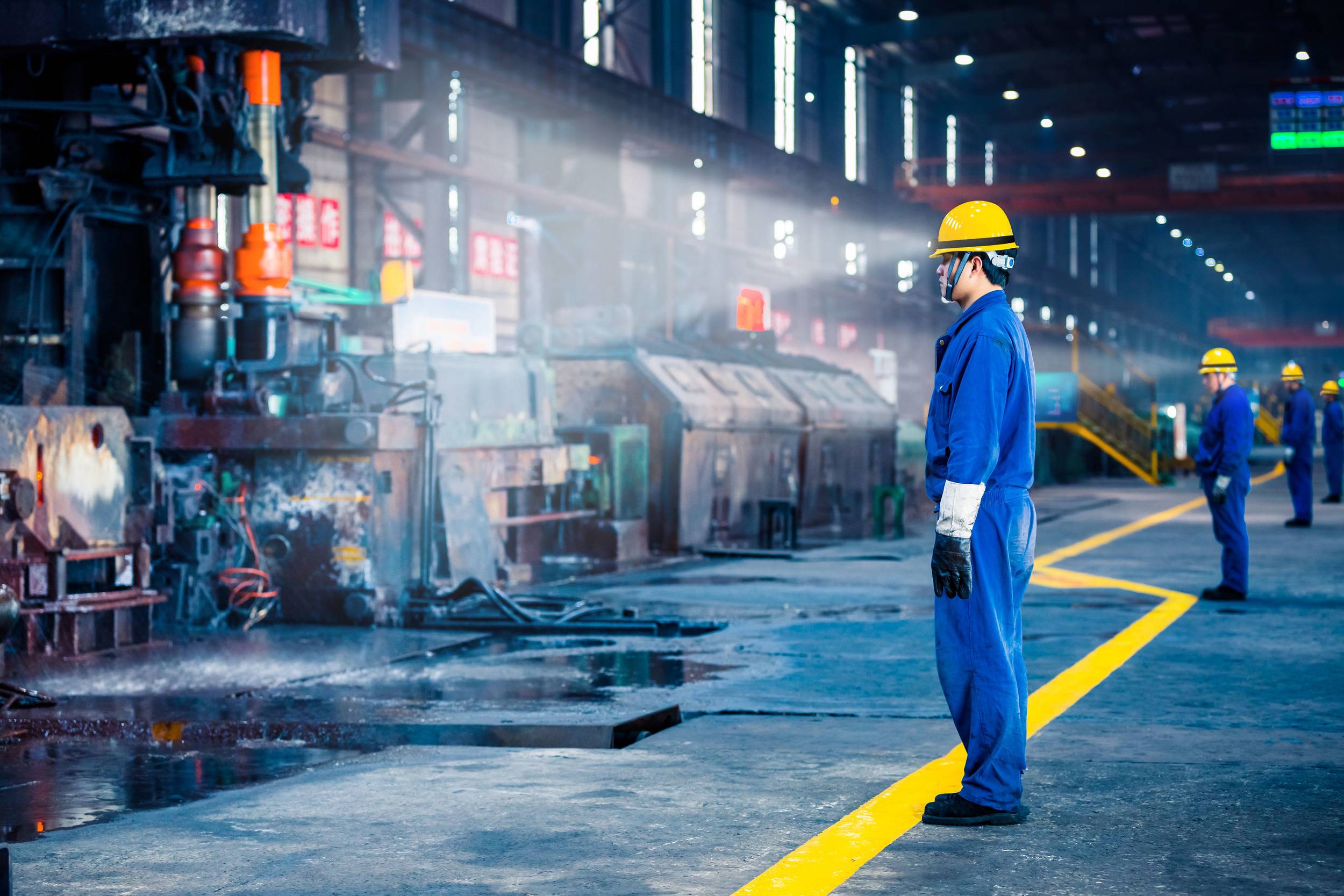 Arbeiter in einer großen Fabrikhalle mit Schutzkleidung und Helmen, die an einer Produktionslinie arbeiten. Industrielle Maschinen im Hintergrund.