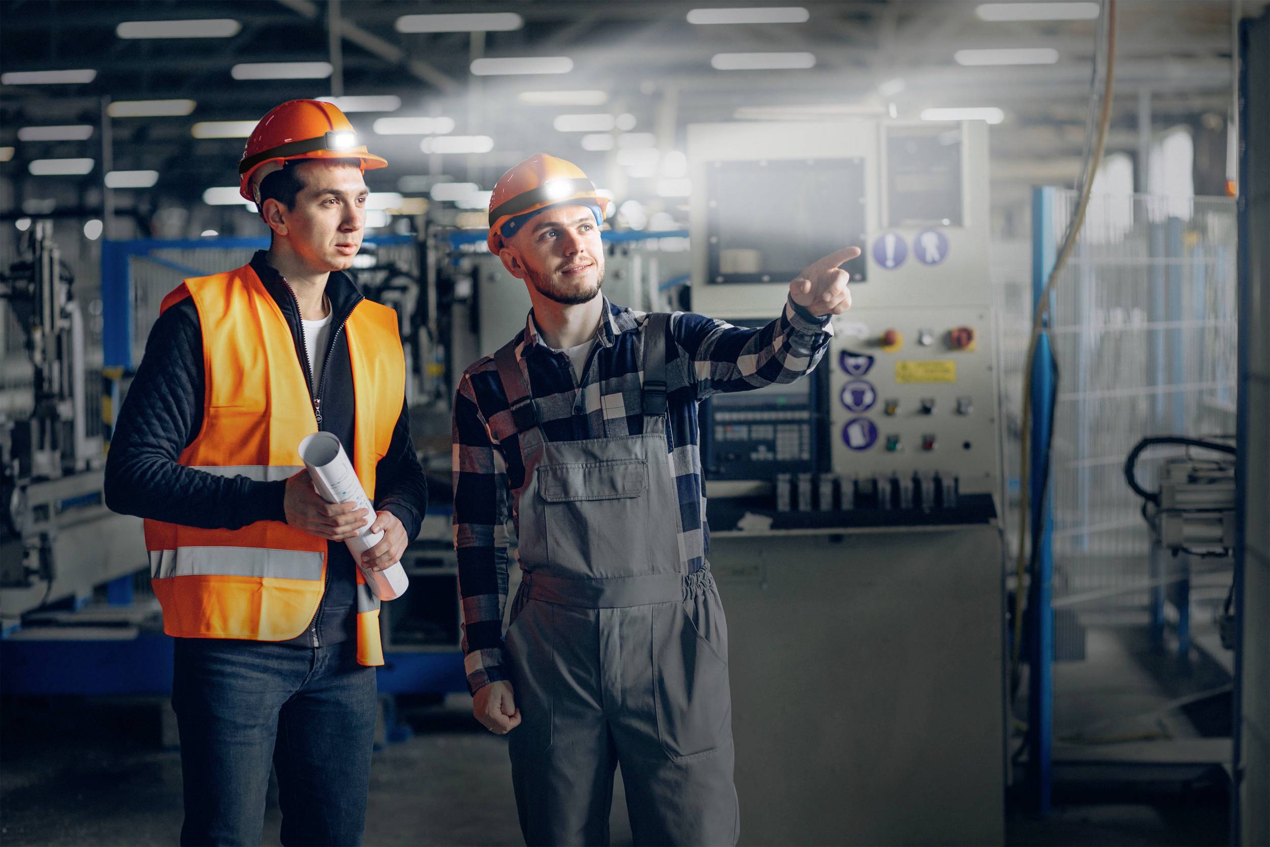 Zwei Arbeiter in Schutzhelmen und Warnwesten stehen in einer Fabrikhalle, einer zeigt auf eine Maschine, der andere hält ein Dokument.