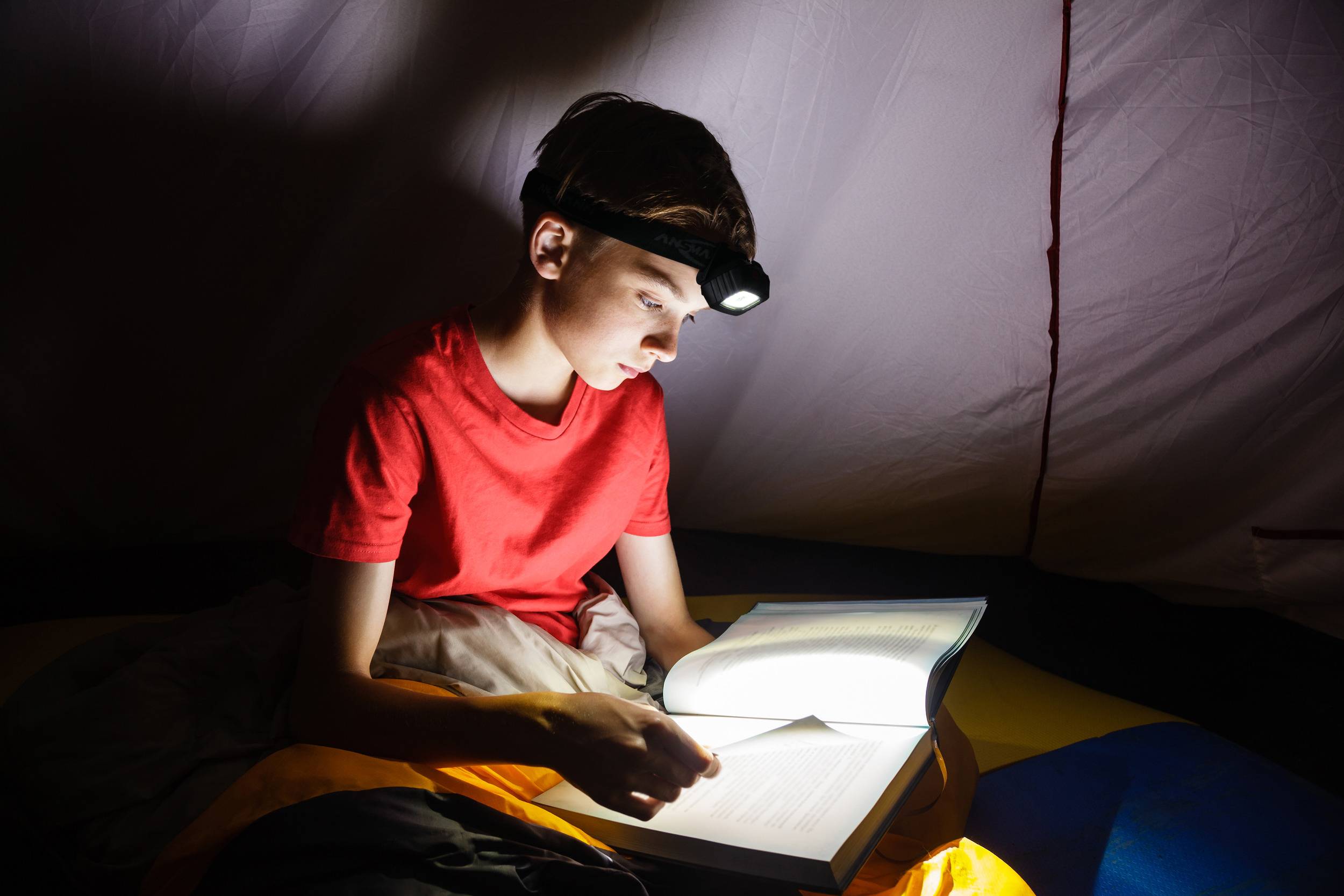 Ein Junge liest nachts unter einer Decke mit Stirnlampe ein Buch, fokussiert und interessiert.