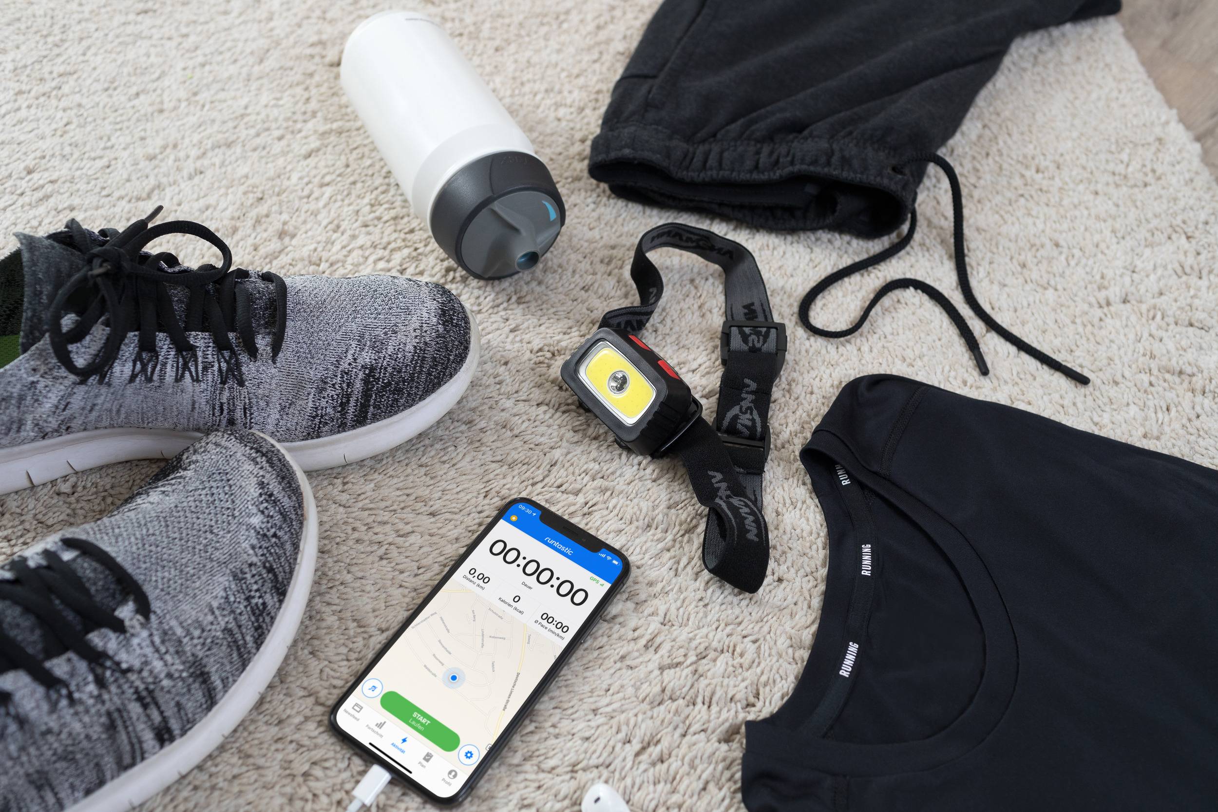 Sportbekleidung und Zubehör liegen auf einem Teppich: Schuhe, Trinkflasche, Kopfhörer, Laufuhr, Trainingshose und ein Smartphone mit offener Fitness-App.