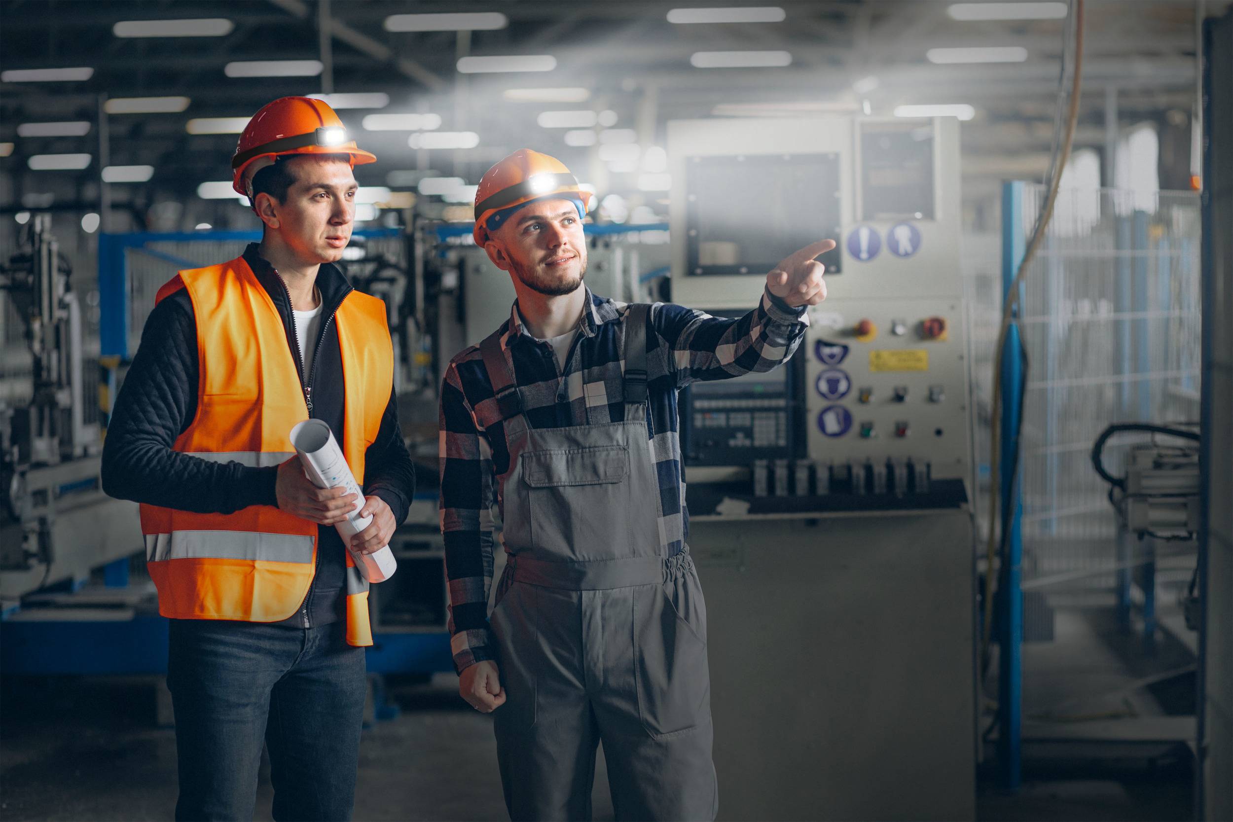 Zwei Arbeiter in Sicherheitskleidung stehen in einer Fabrikhalle. Einer hält Baupläne, der andere zeigt auf eine Maschine im Hintergrund.
